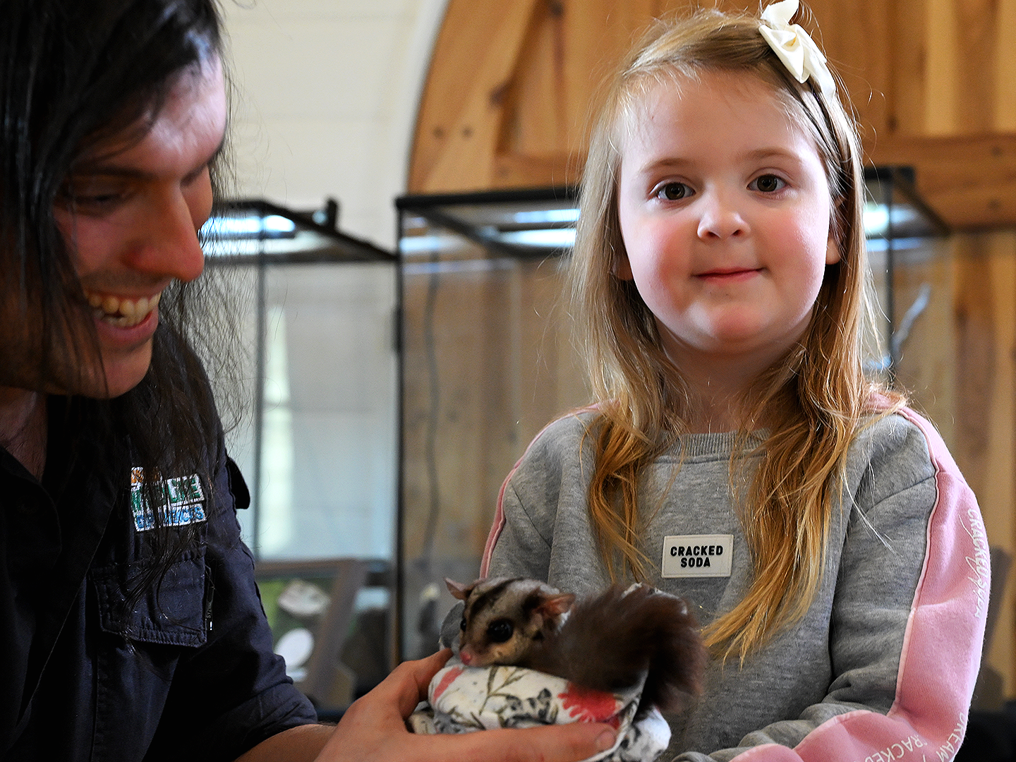 A young girl with long blonde hair holding a sugar glider, wrapped in a blanket, while a man with dark hair smiles beside her. There are animal enclosures in the background.
