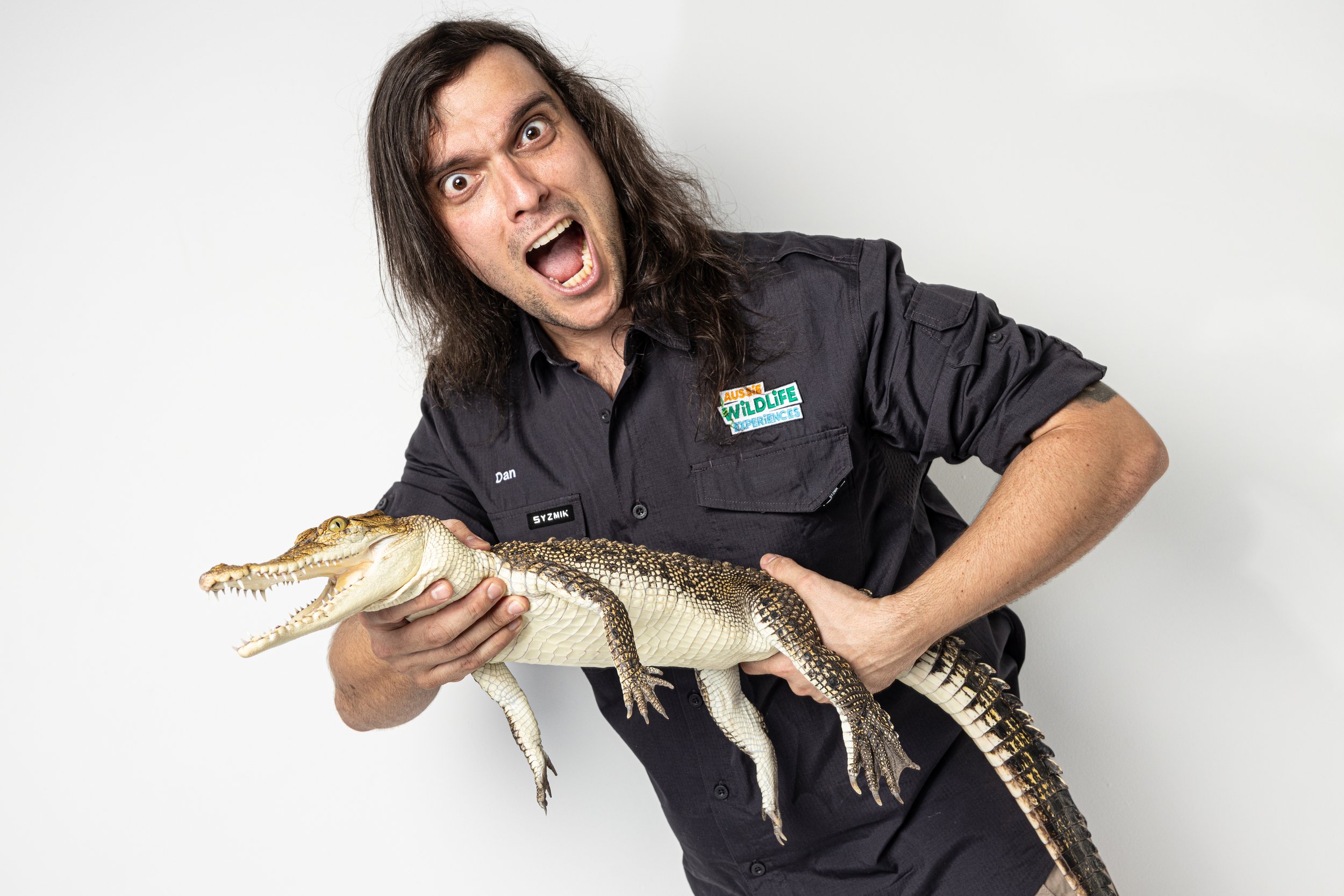 A man with long dark hair wearing a black shirt with a 'Aussie Wildlife Experiences' patch on it, holding a small crocodile.