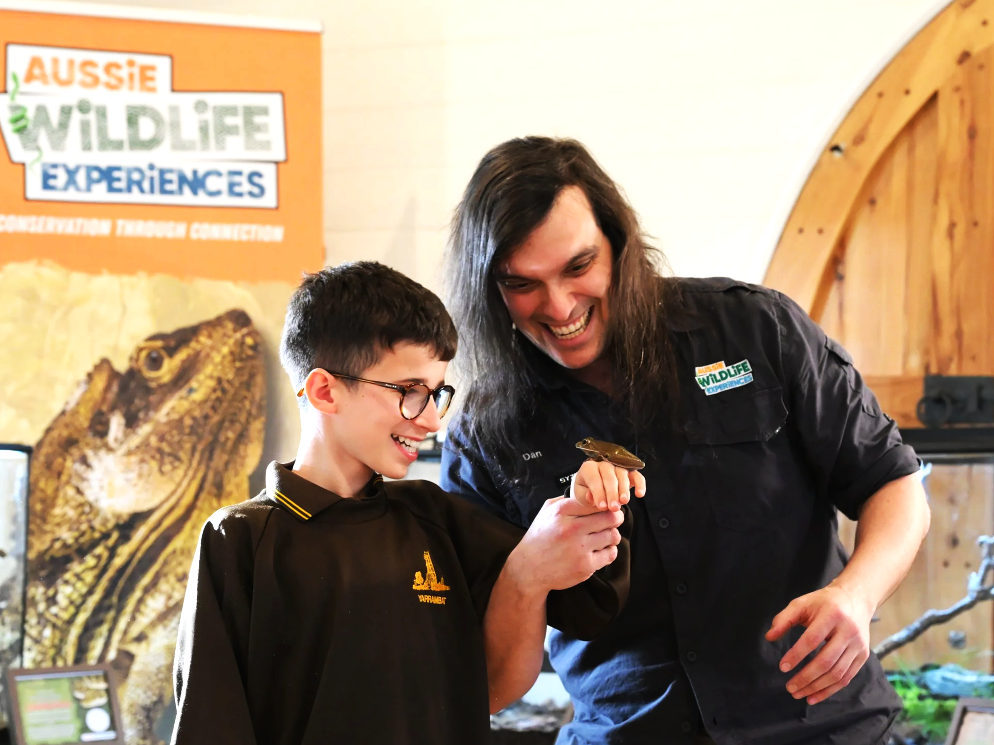 A young boy and a man with long hair holding a frog, at a wildlife experience event with a sign in the background that reads "Aussie Wildlife Experiences" and a picture of a frilled-neck lizard.