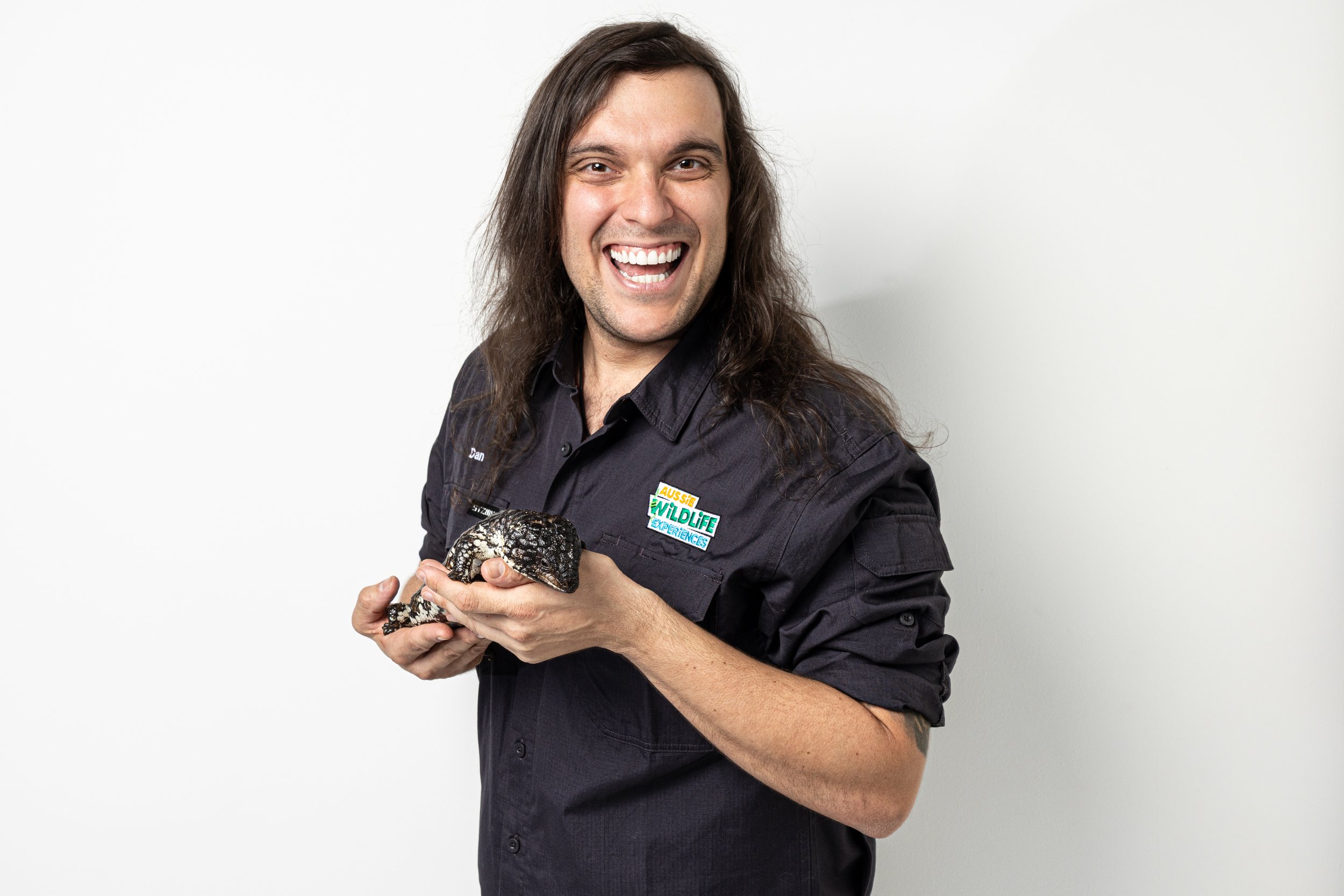 A man with long brown hair smiling broadly while holding a shingleback lizard. He is wearing a black shirt with a patch that reads 'Aussie Wildlife Experiences'.