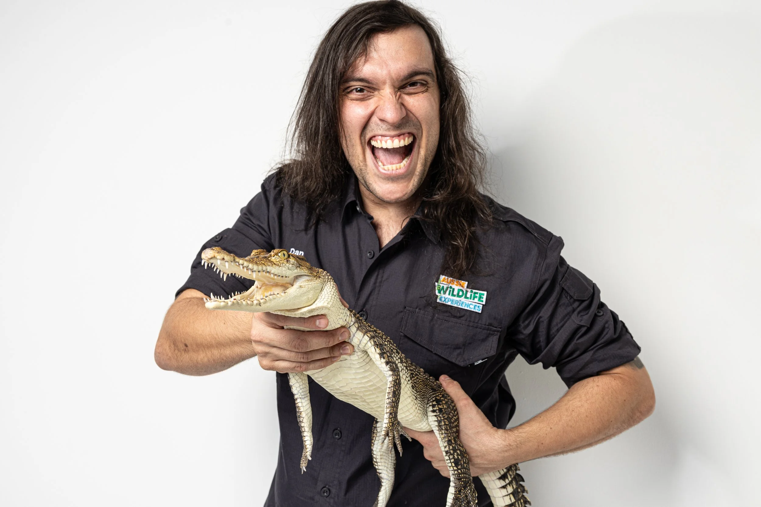 A man with long dark hair, wearing a black shirt, is smiling widely and holding a small crocodile in his hands. The background is plain white.