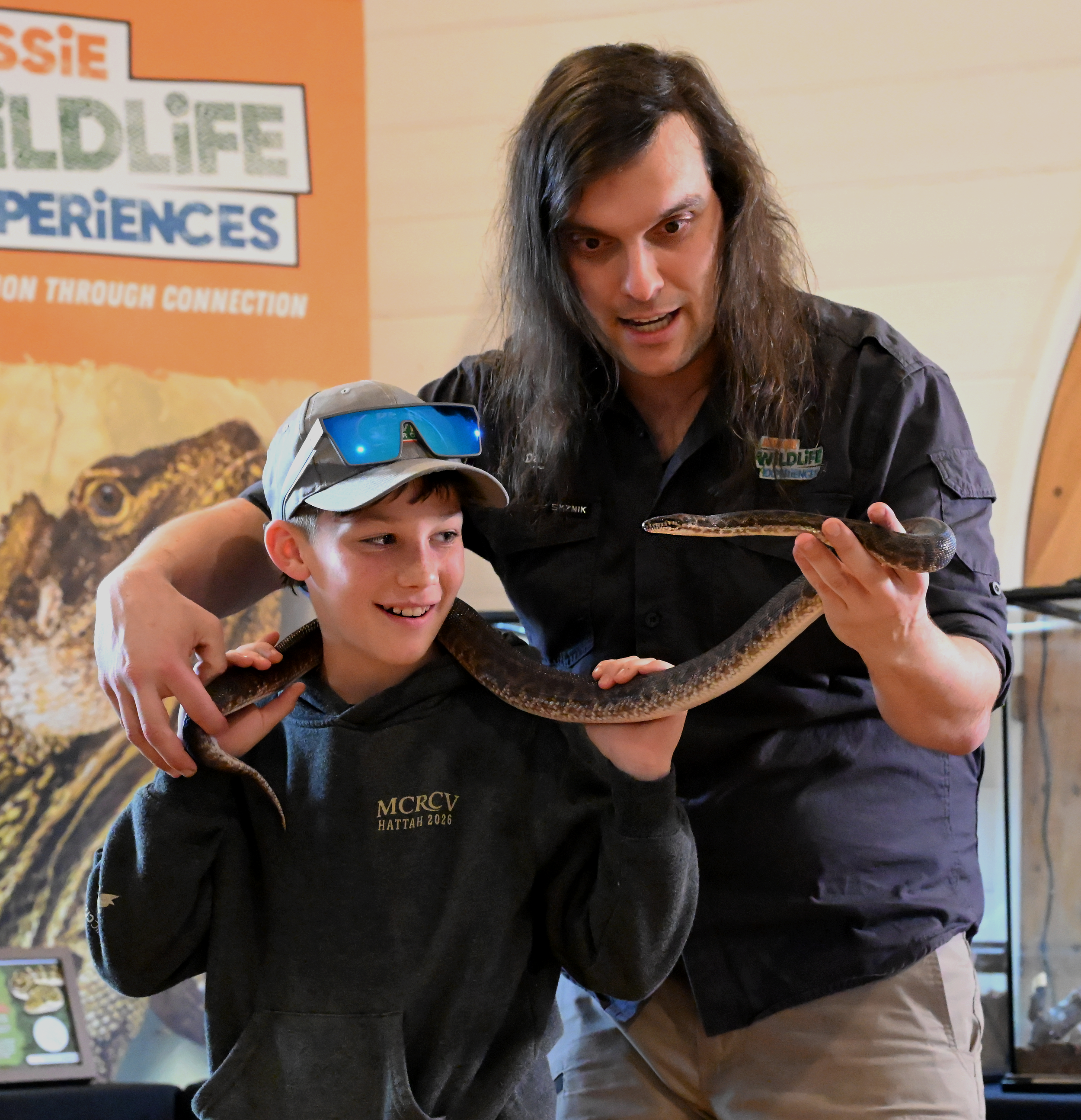 A man and a boy holding a snake at a wildlife exhibit. The man wears a black shirt with a wildlife logo, and the boy wears a cap with sunglasses on top. There is a colorful background with a sign that reads "Aussie Wildlife Experiences."