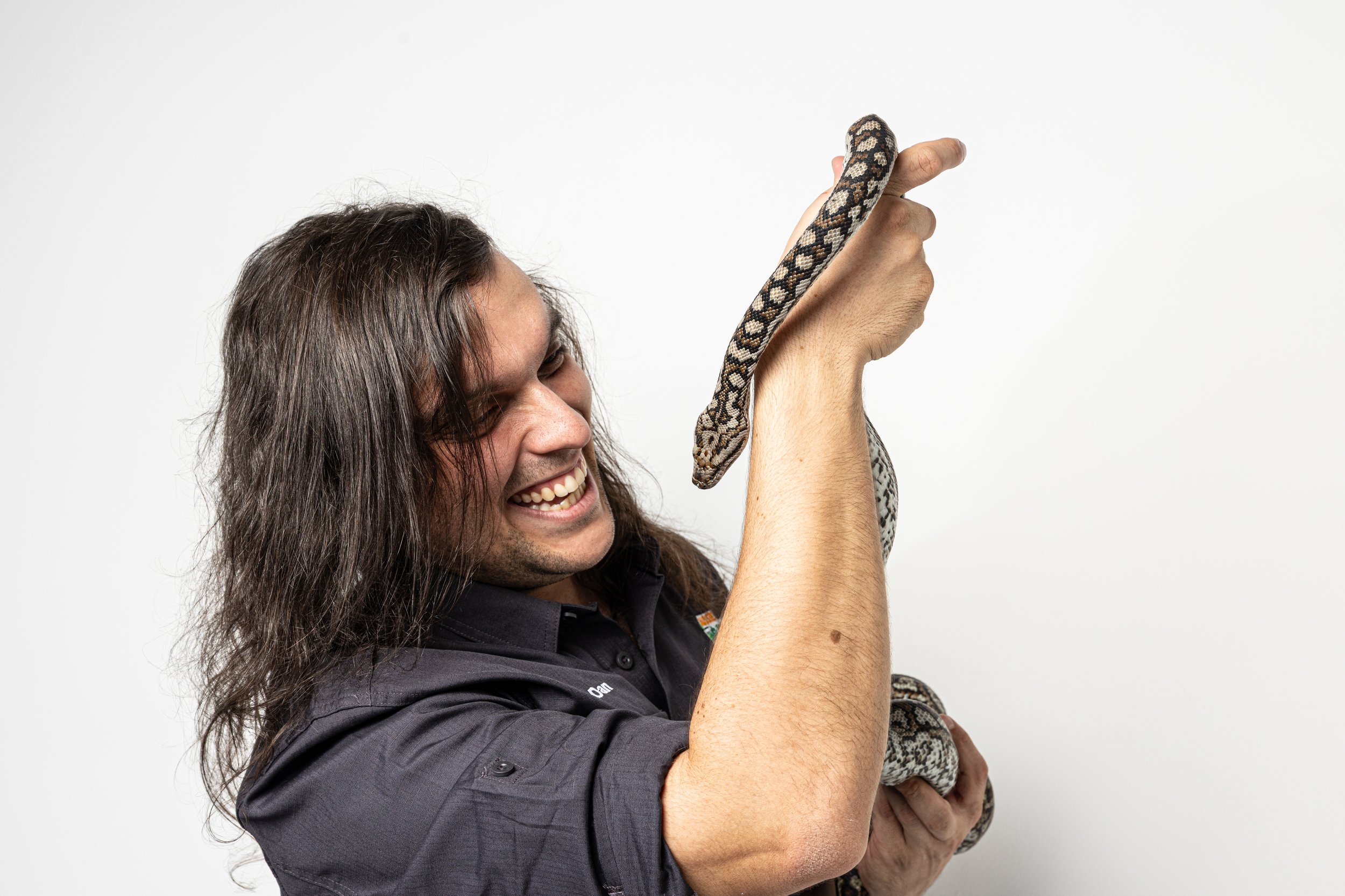 A man with long, dark hair smiling and wearing a black collared shirt is holding a snake up with his right hand, while his left arm is bent and supporting his right elbow, against a plain white background.