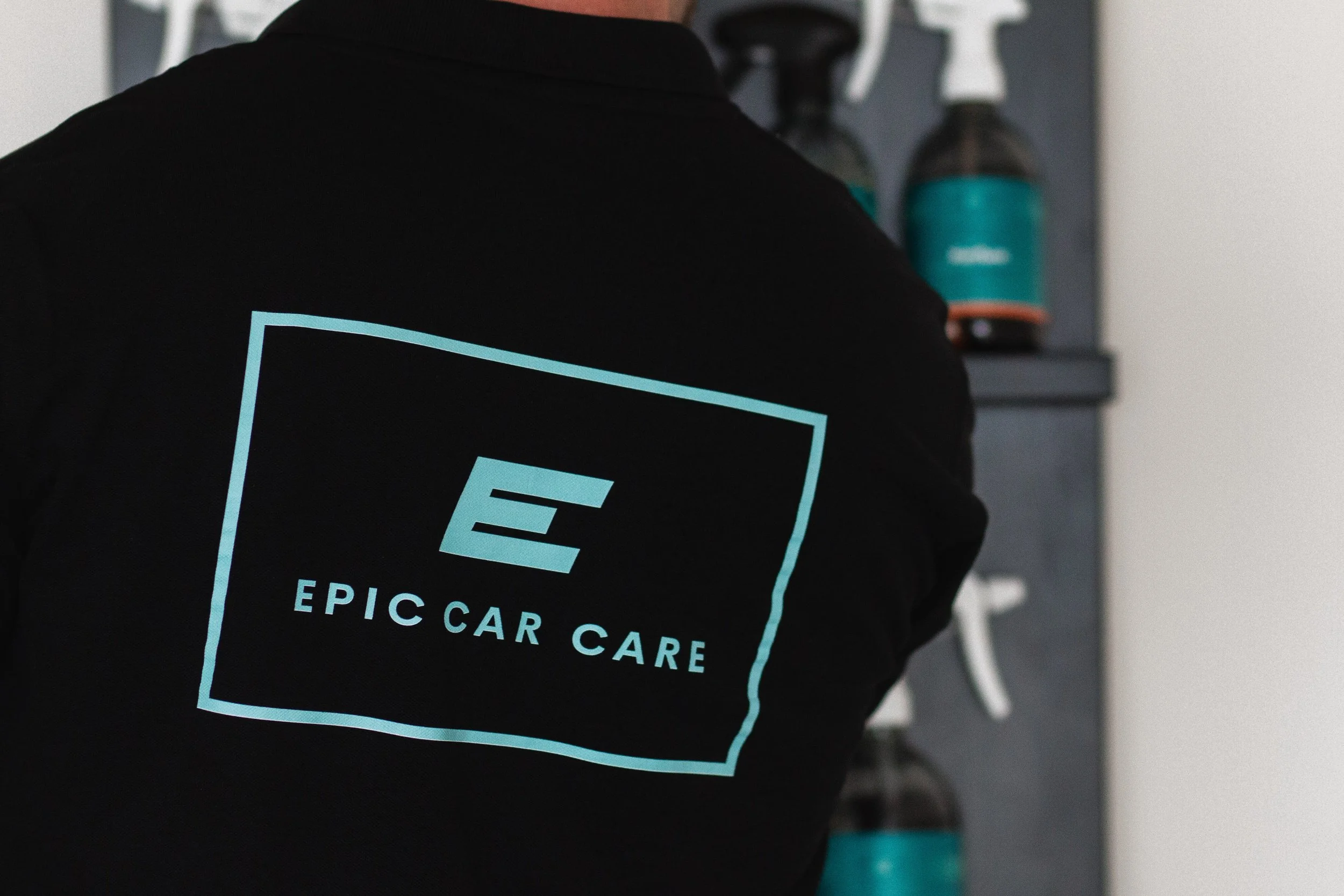 Close-up van een persoon die een zwart kledingstuk draagt met een logo van EPIC CAR CARE erop, met op de achtergrond een muur en enkele flesjes, mogelijk shampoo of schoonmaakmiddel.