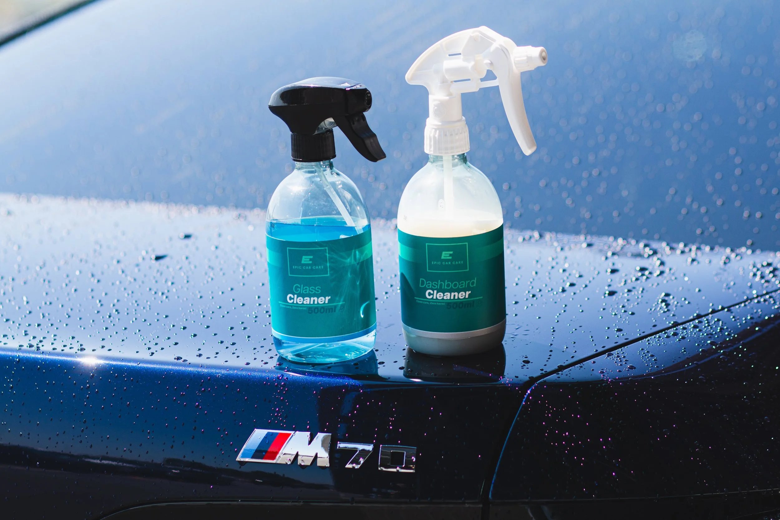 Twee sprayflessen met autoshampoo en interieurreiniger op de motorkap van een zwarte BMW M7, met regen en waterdruppels op de auto en een badge van BMW M.