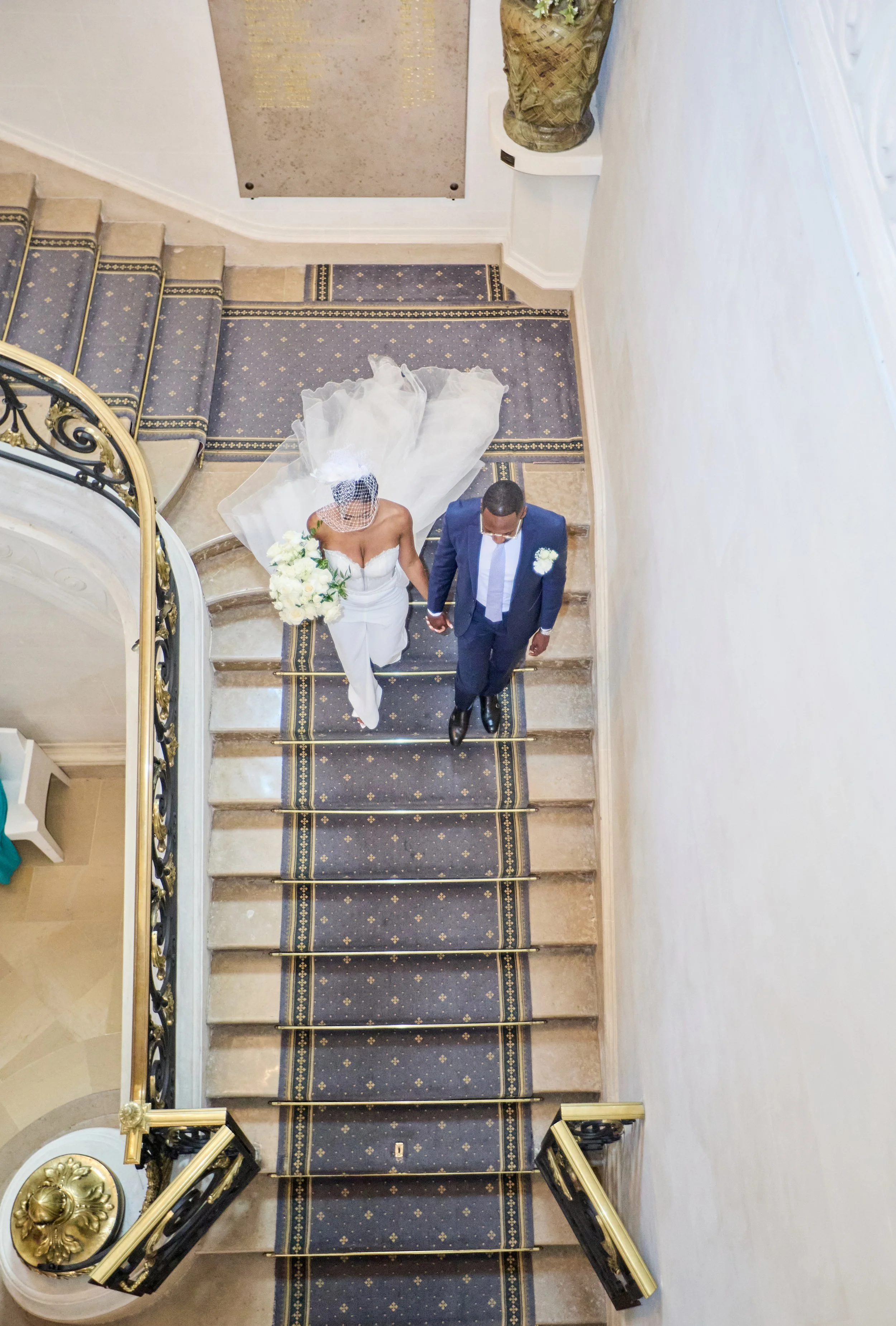 Une personne portant une robe de mariage blanche et un voile, accompagnée d'un homme en costume, marche dans un bâtiment élégant.