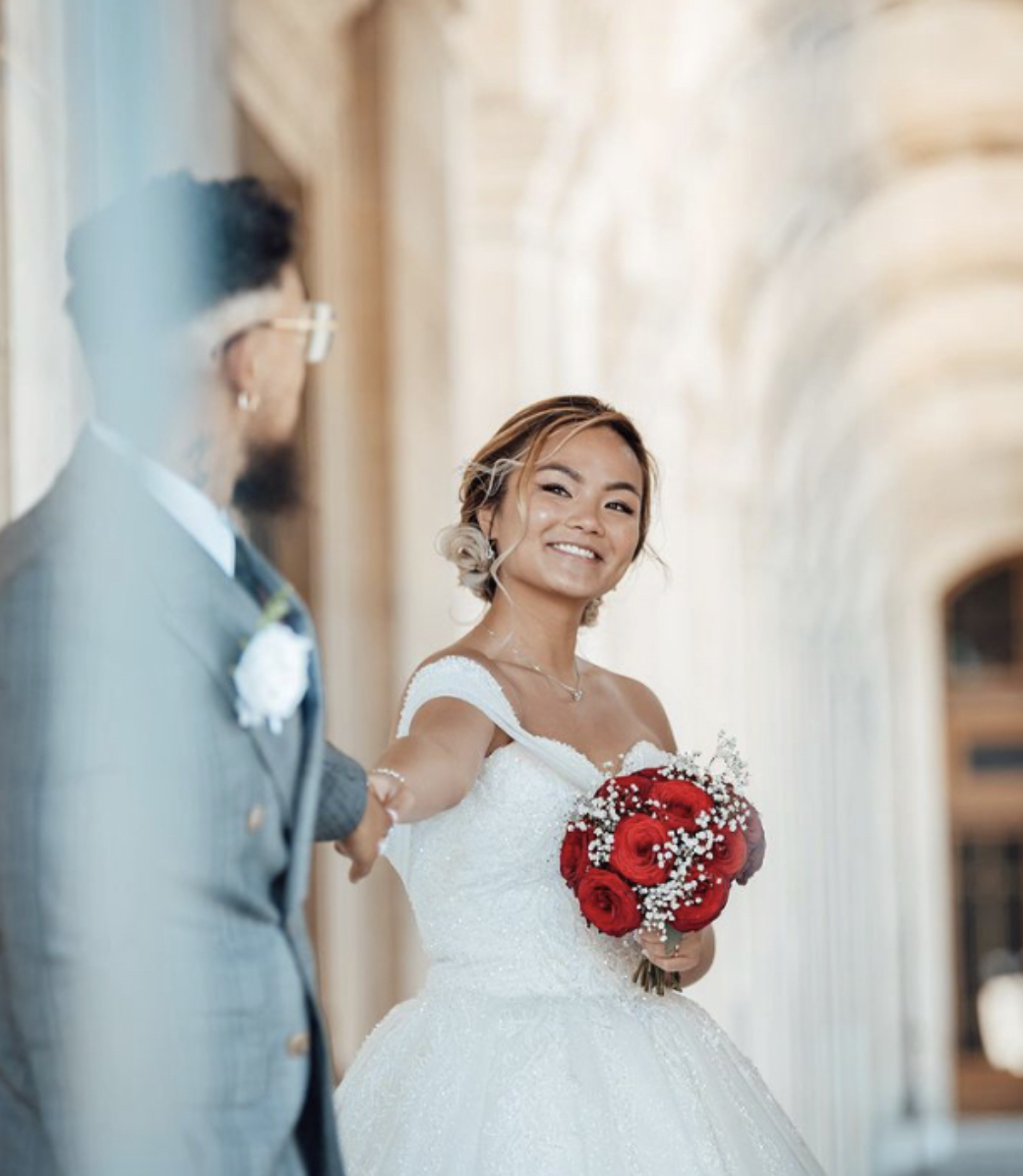 Une femme en robe de mariée tenant un bouquet de roses rouges sourit, tandis qu'un homme en costume de mariage lui serre la main dans un couloir élégant.