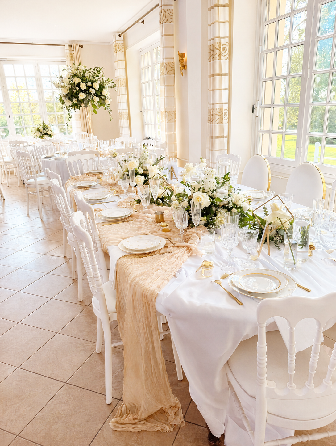 Salle de réception décorée pour un mariage avec une grande table longue recouverte de nappe blanche, ornée de centres de table avec des fleurs blanches et de la verdure, des assiettes en porcelaine blanche avec des assiettes plates et des couverts dorés, des verres à vin et à eau, sous une lumière naturelle provenant de grandes fenêtres. La salle a des murs clairs, des rideaux assortis et un sol en carrelage beige.