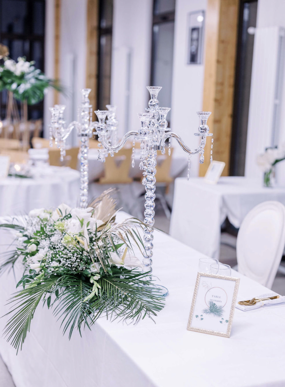 Table décorée avec un chandelier en cristaux et une composition florale composée de fleurs blanches et de feuilles vertes, dans un cadre élégant de style mariage ou événement spécial.