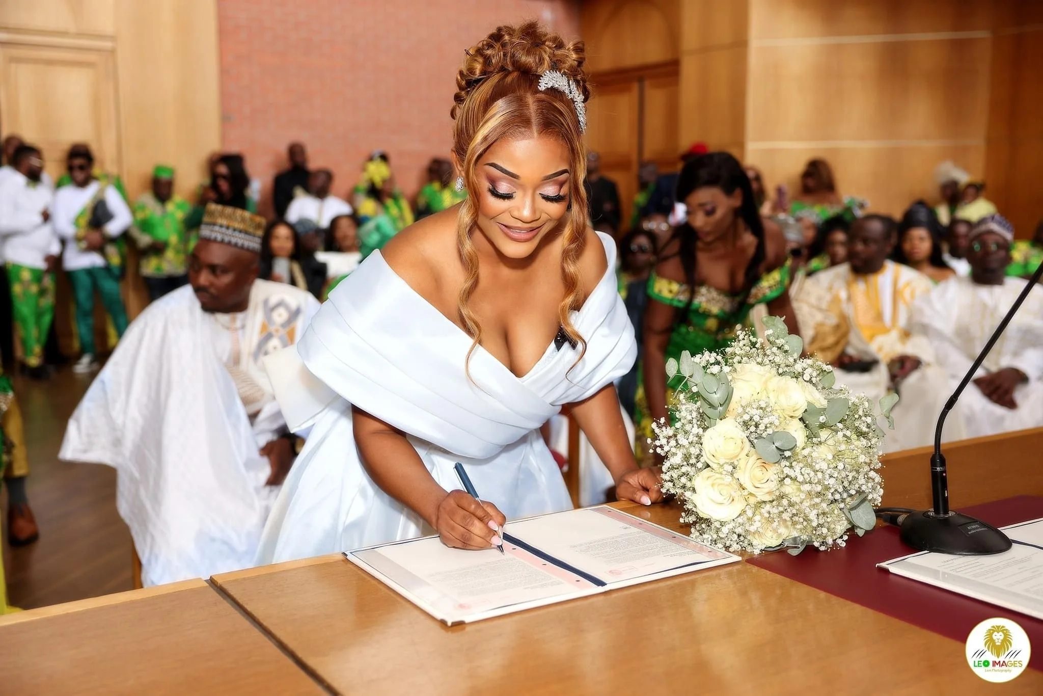 Une femme en robe blanche signe un document lors d'une cérémonie de mariage, entourée de personnes en tenues traditionnelles colorées dans une salle en bois.