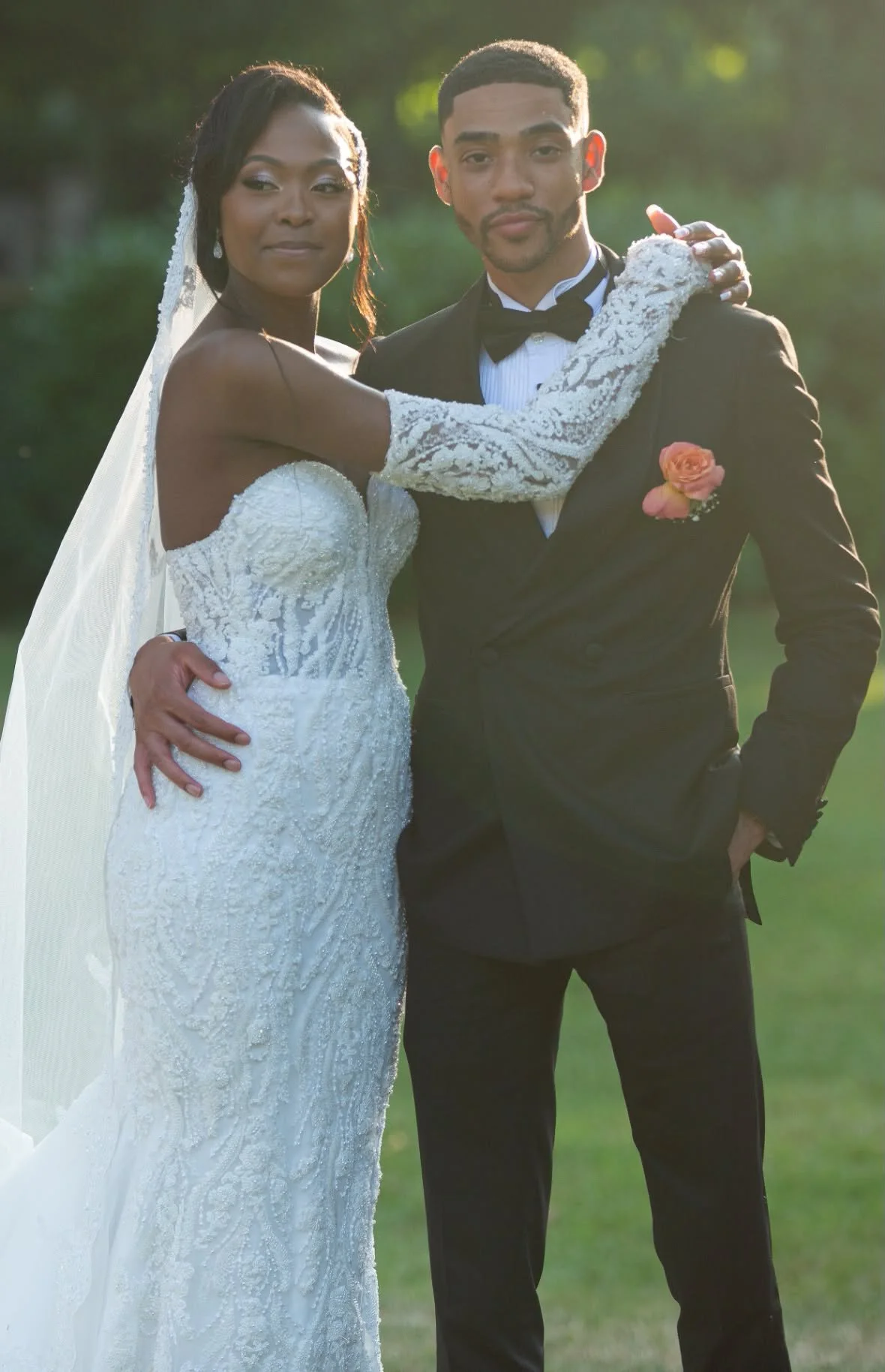 Un couple en tenue de mariage posant à l'extérieur, la femme porte une robe de mariée blanche avec des manches longues en dentelle, et l'homme porte un smoking noir avec un nœud papillon, un bouton de rose sur la veste, lors d'une séance photo en ple