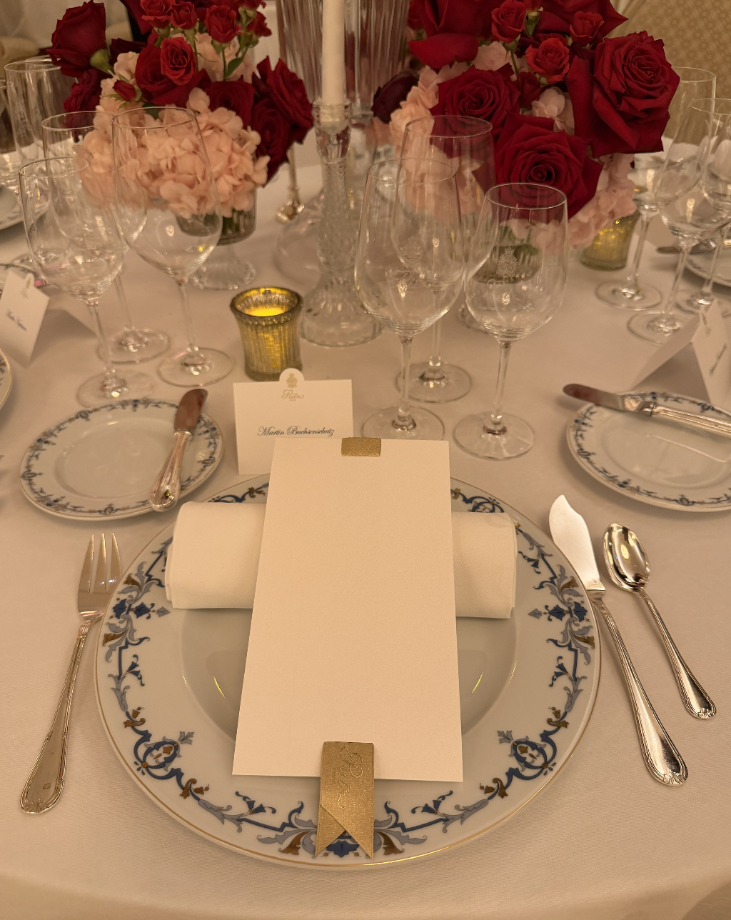 Table de dîner élégante avec des fleurs rouges et roses, des verres à vin, des assiettes en porcelaine décorées, et des couverts en argent, prête pour un repas sophistiqué.