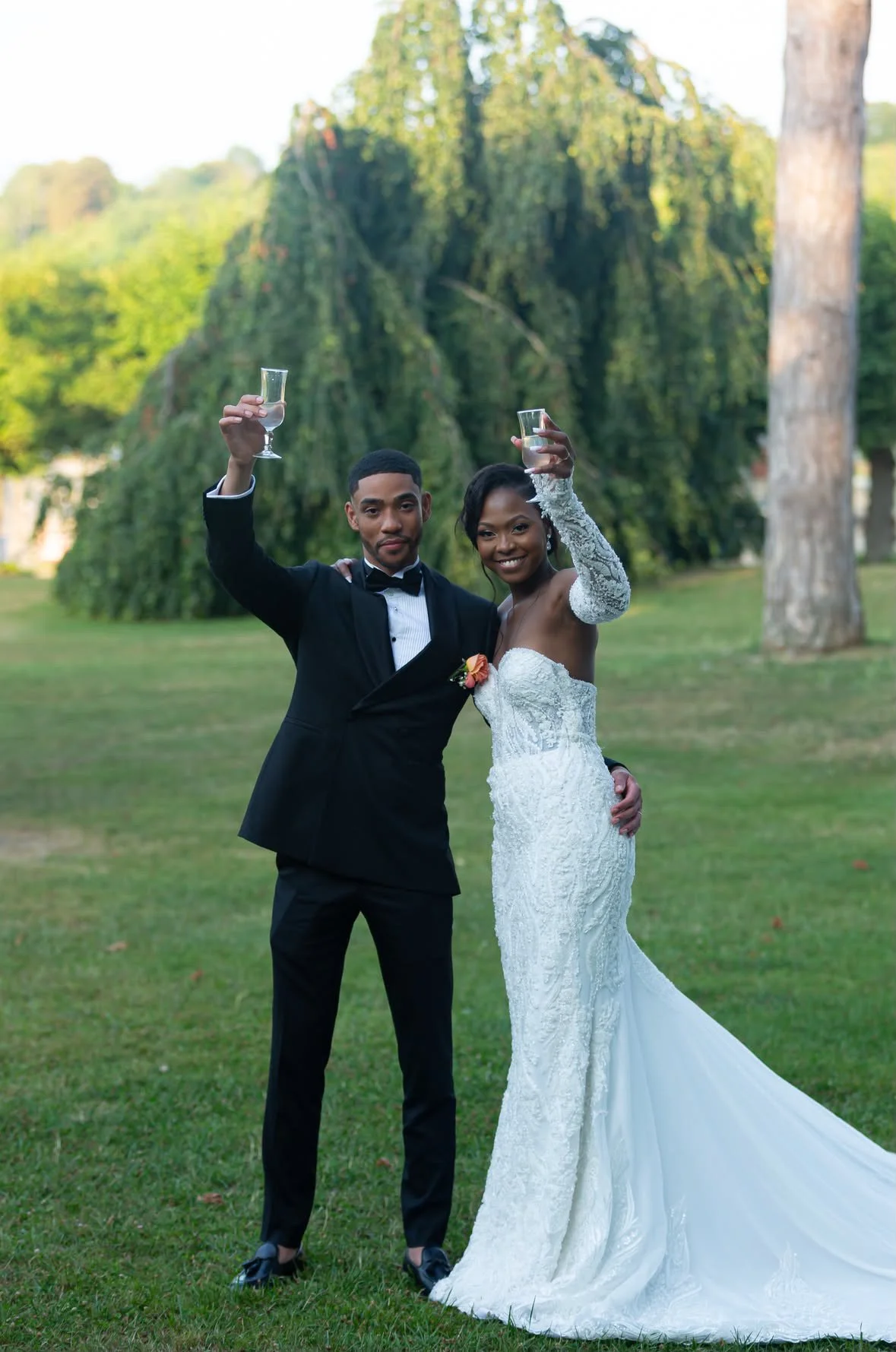 Un couple en costume de mariage levant des verres pour faire un toast dans un parc avec des arbres en arrière-plan.