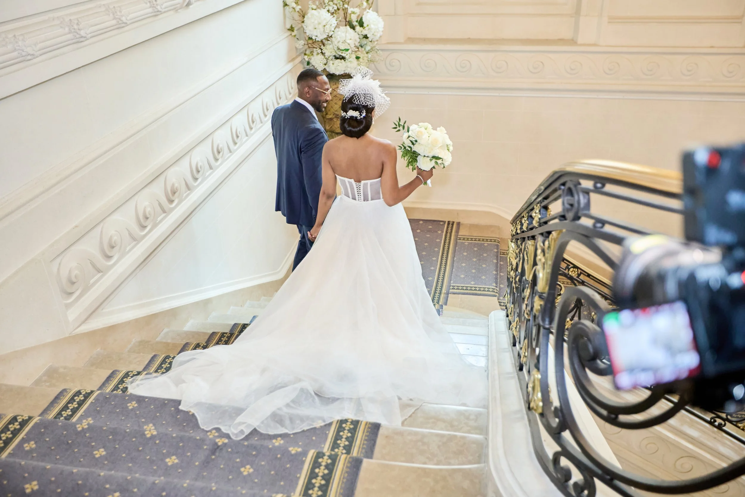 Un couple de mariés marchant dans un escalier dans une salle de mariage, la femme portant une robe de mariée blanche avec un bouquet de fleurs, l'homme en costume bleu. La scène est dans un décor élégant avec des fleurs blanches en arrière-plan.