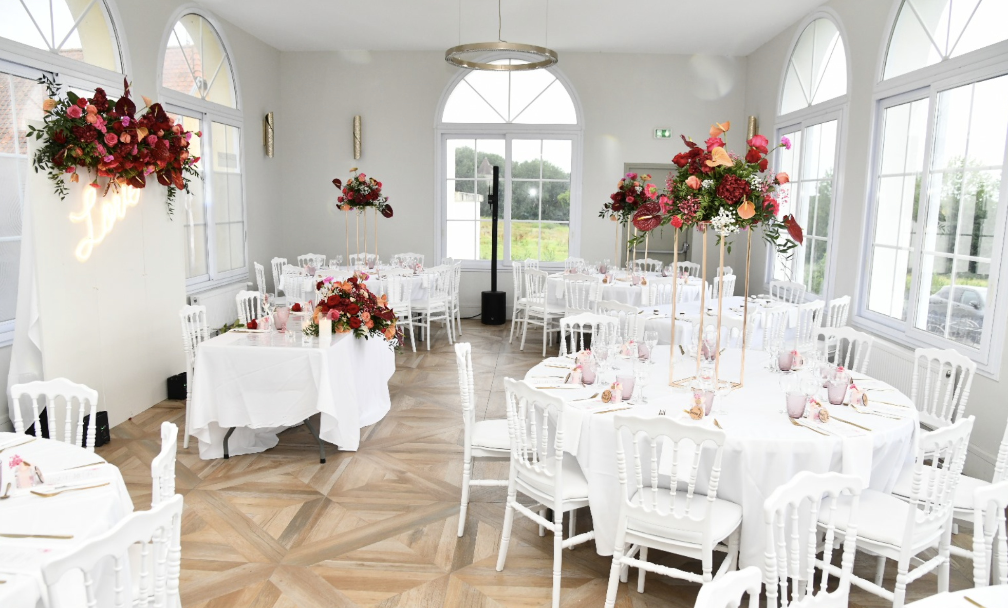 Salle de réception décorée pour un mariage avec tables rondes, fleurs colorées en centre de table, et grandes fenêtres laissant entrer la lumière