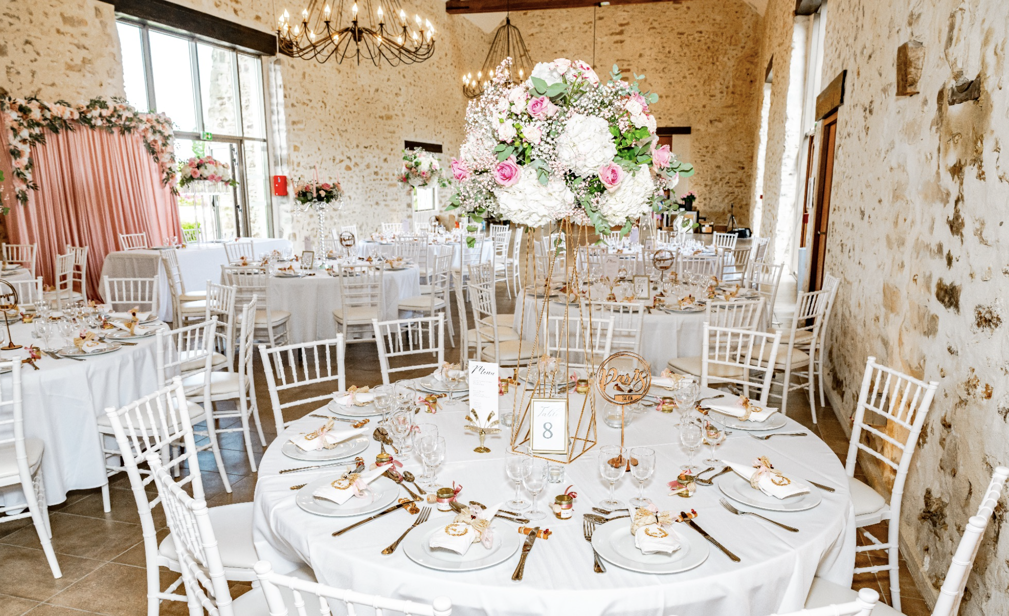 Salle de mariage décorée avec des tables rondes vêtues de nappes blanches, ornées d'une Grande composition florale centrale composée de roses, hortensias et feuillage; la pièce a des murs en pierre et des chandeliers suspendus au plafond.