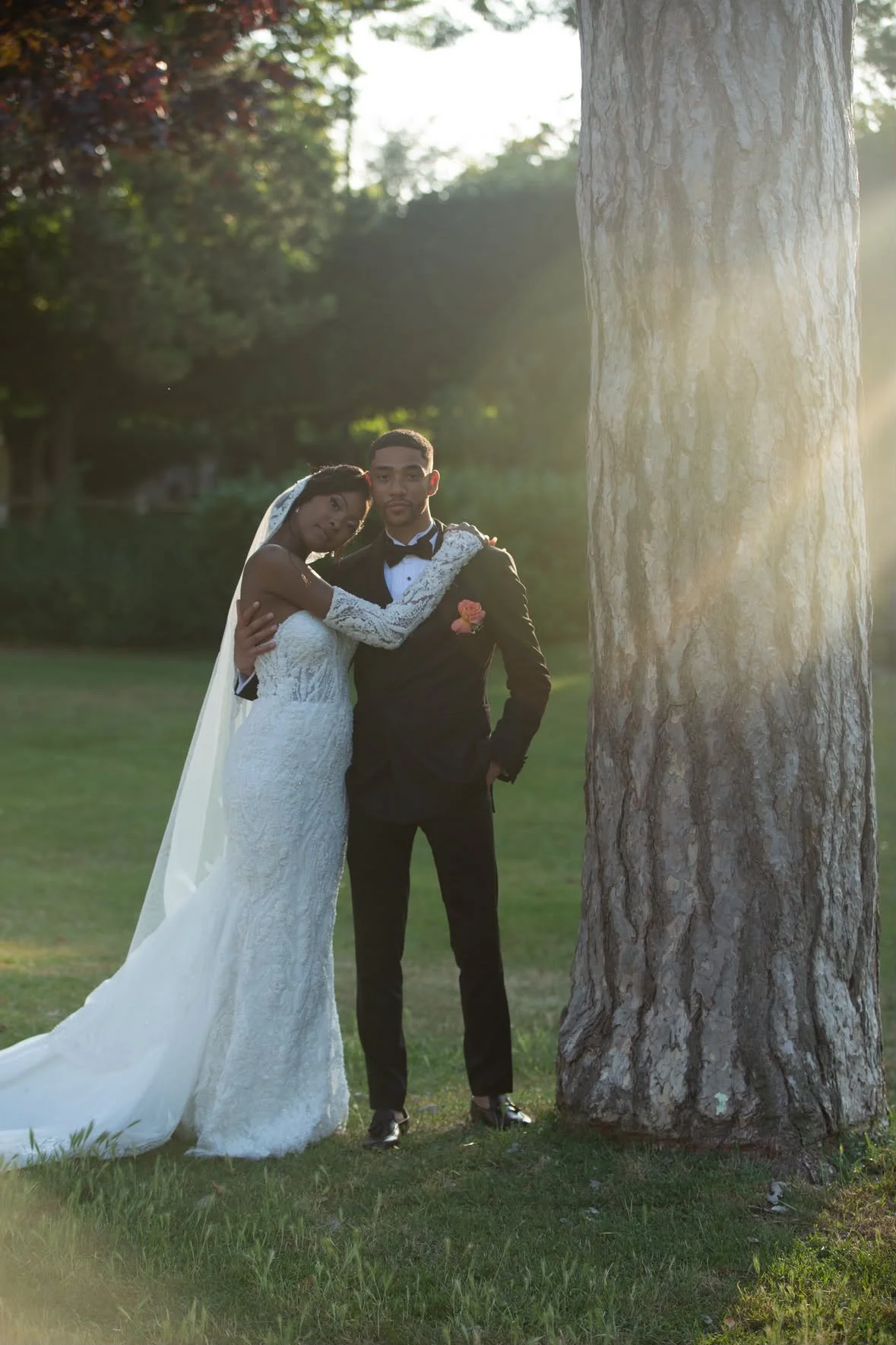 Un couple en mariée et marié pose sous un arbre lors d'une séance photo en plein air, en fin d'après-midi ou en début de soirée.