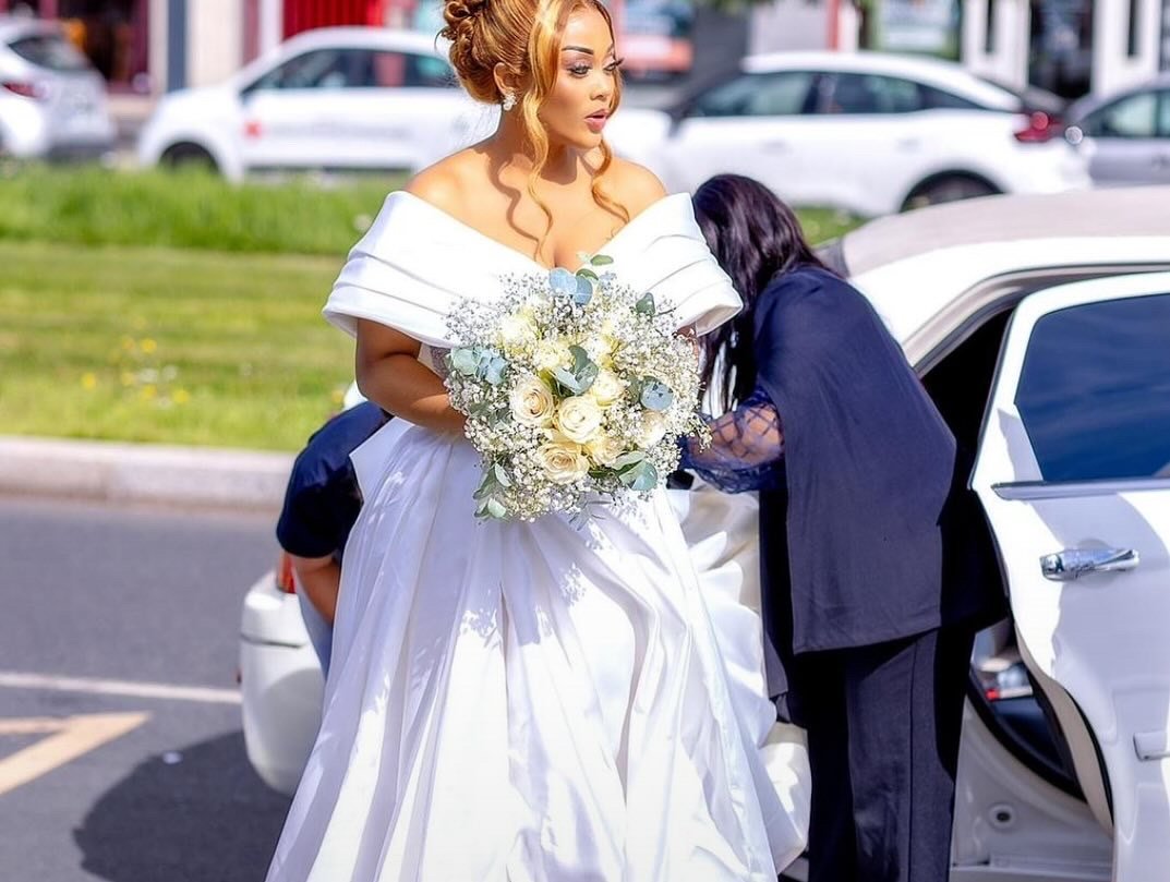 Une femme en robe de mariée tenant un bouquet de fleurs blancs, un homme en costume sombre l'aide à entrer dans une voiture blanche.