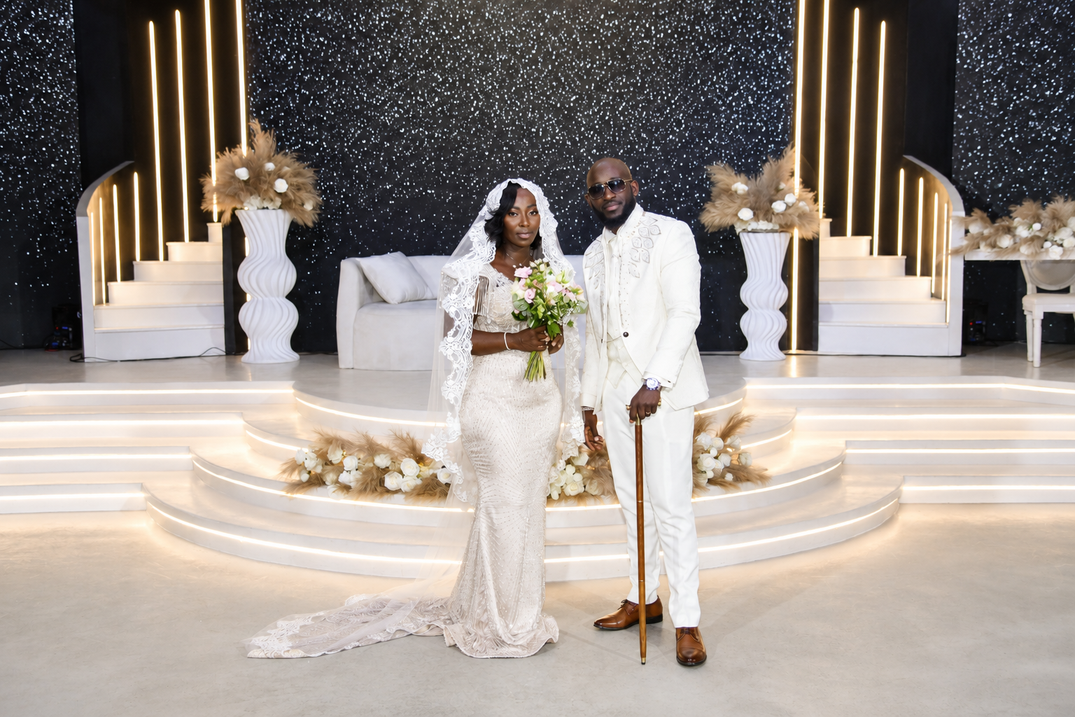 Un couple de mariés en costume blanc posant lors de leur mariage, avec décor sophistiqué comprenant des escaliers, des vases blancs avec des fleurs et un fond étoilé lumineux.