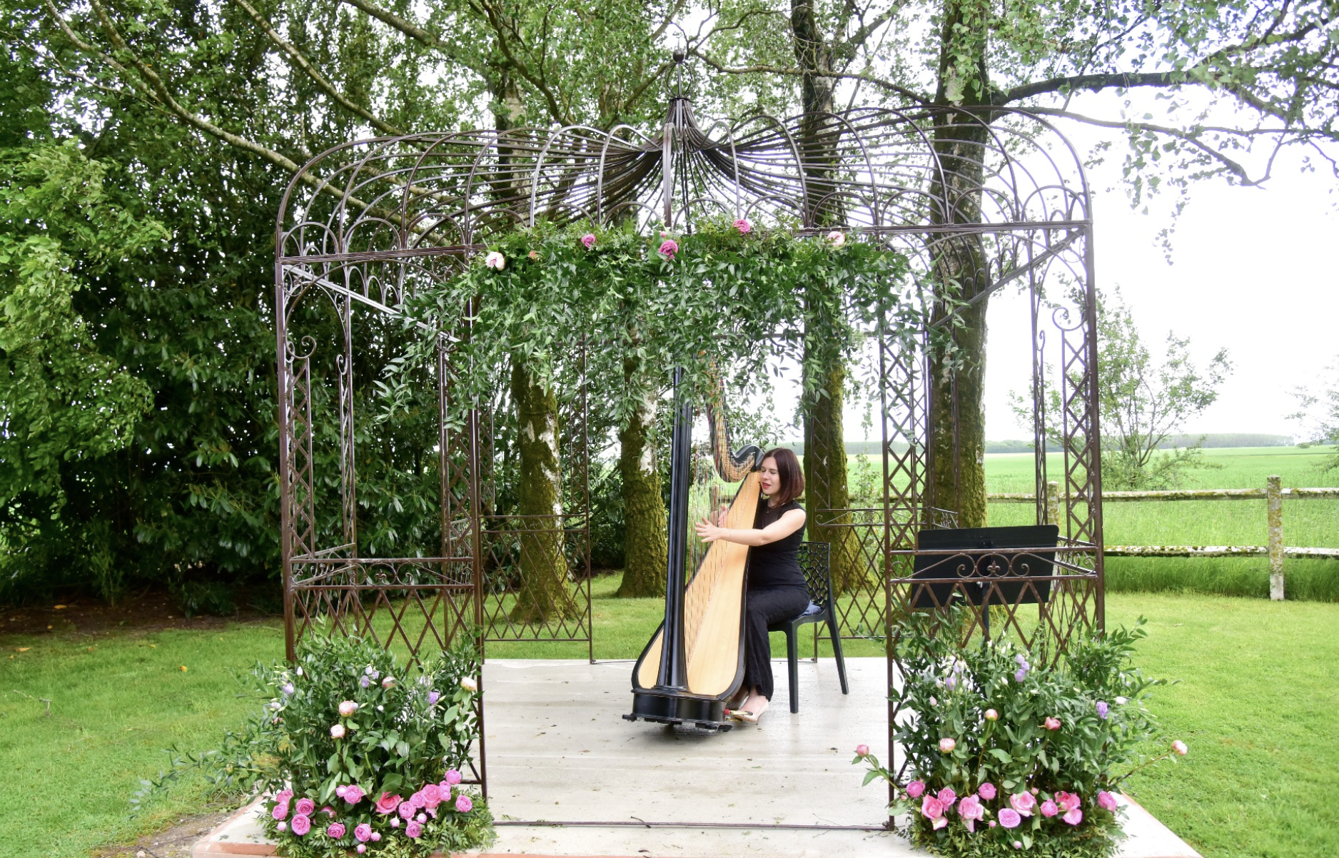 Femme jouant de la harpe dans un kiosque en fer forgé, entouré de fleurs roses, avec un paysage rural en arrière-plan.
