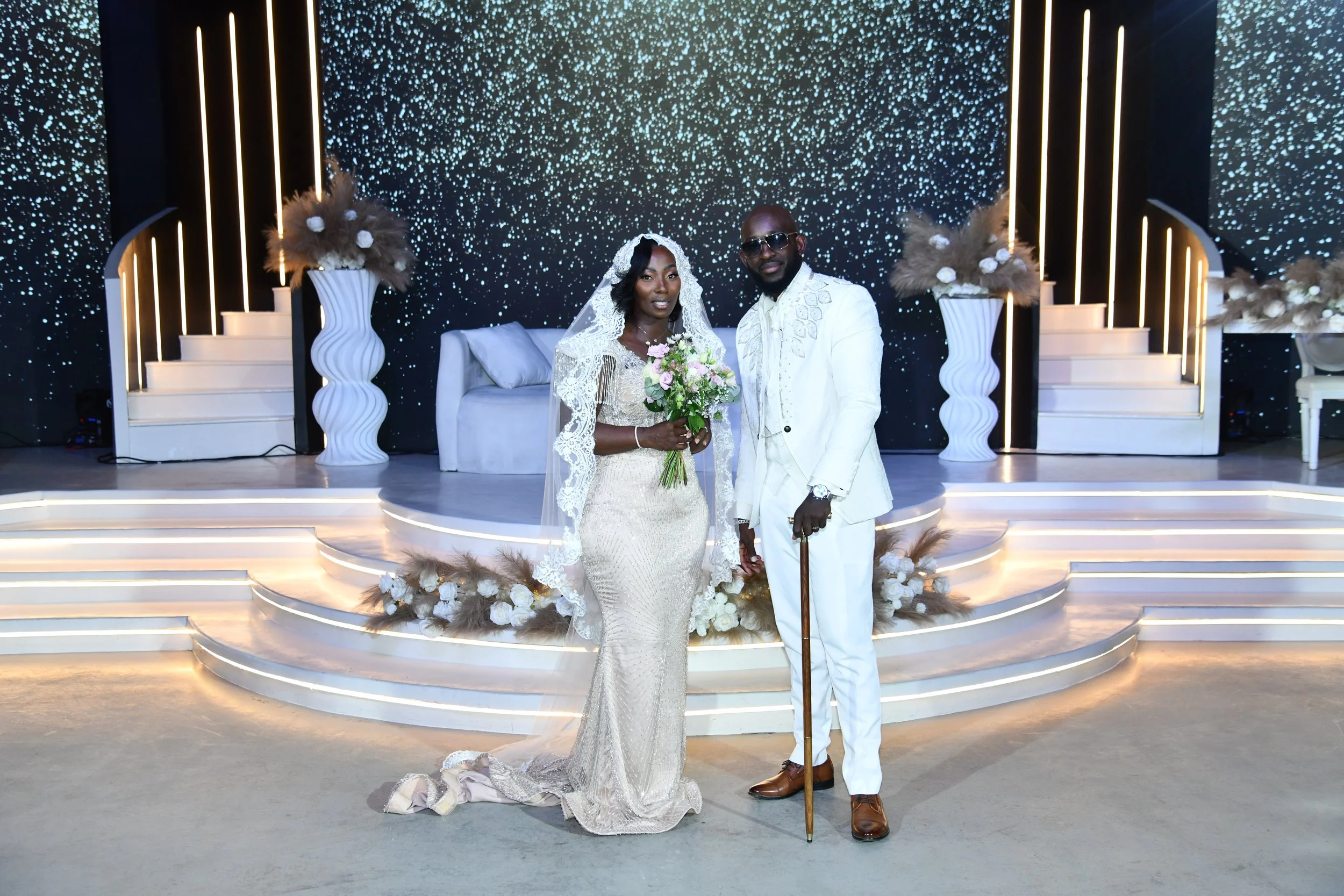 Un couple en mariage sur une scène décorative moderne avec des vases de fleurs et un fond étoilé, vêtus de tenues de mariage, la femme porte une robe blanche avec un voile, et l'homme porte un costume blanc avec des lunettes de soleil.