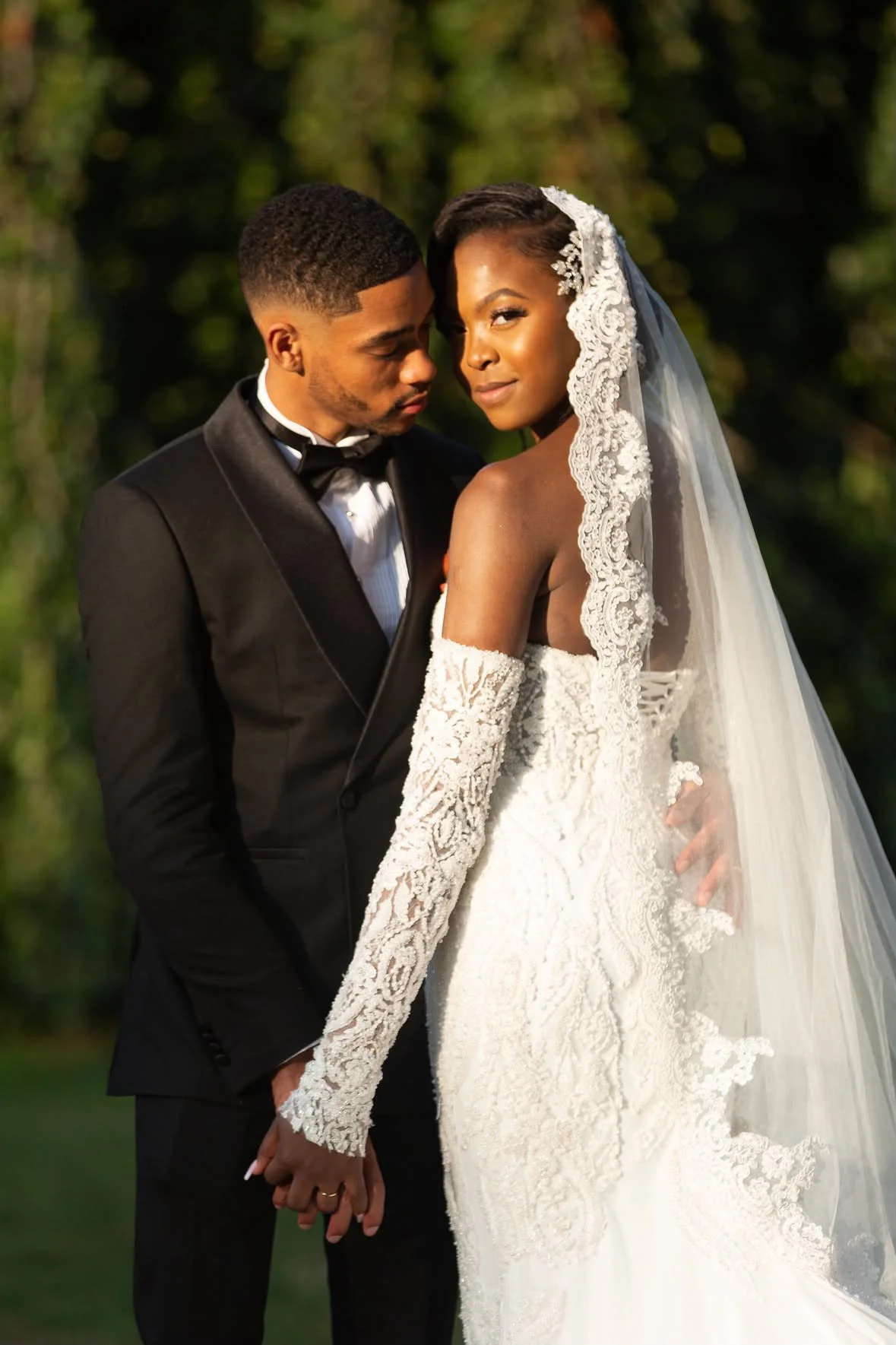 Un couple en tenue de mariage, la femme porte une robe blanche avec des broderies en dentelle, un voile et des mitaines longues en dentelle, l'homme porte un costume noir avec une chemise blanche et un noeud papillon, ils se tiennent par la main, dan