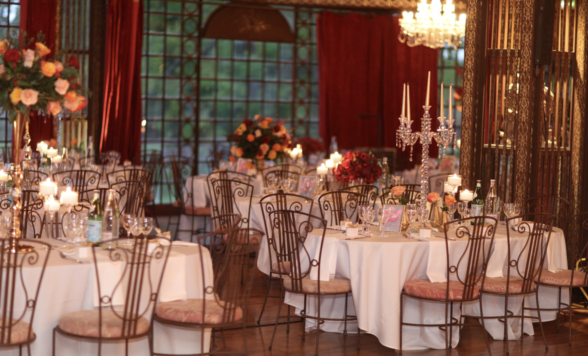 Salle de réception élégante avec des tables rondes couvertes de nappes blanches, décorées de bouquets de fleurs et de bougies, ambiance chaleureuse avec éclairage chandeliers, grands fenêtres en verre et rideaux rouges.