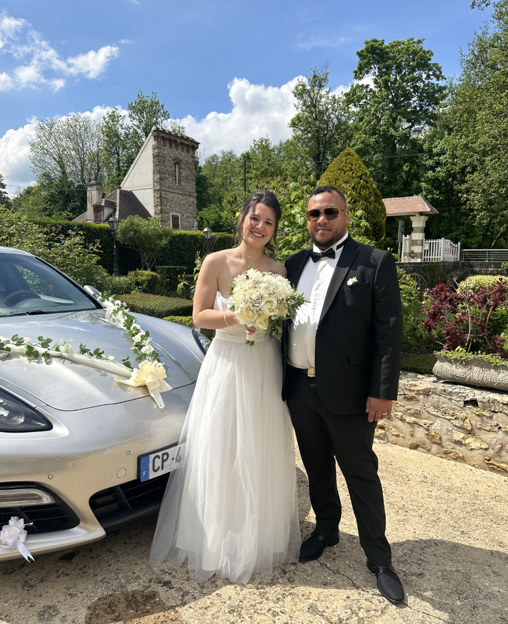 Un couple de mariés posant devant une voiture décorée avec des rubans et fleurs, en plein air, dans un jardin ensoleillé.