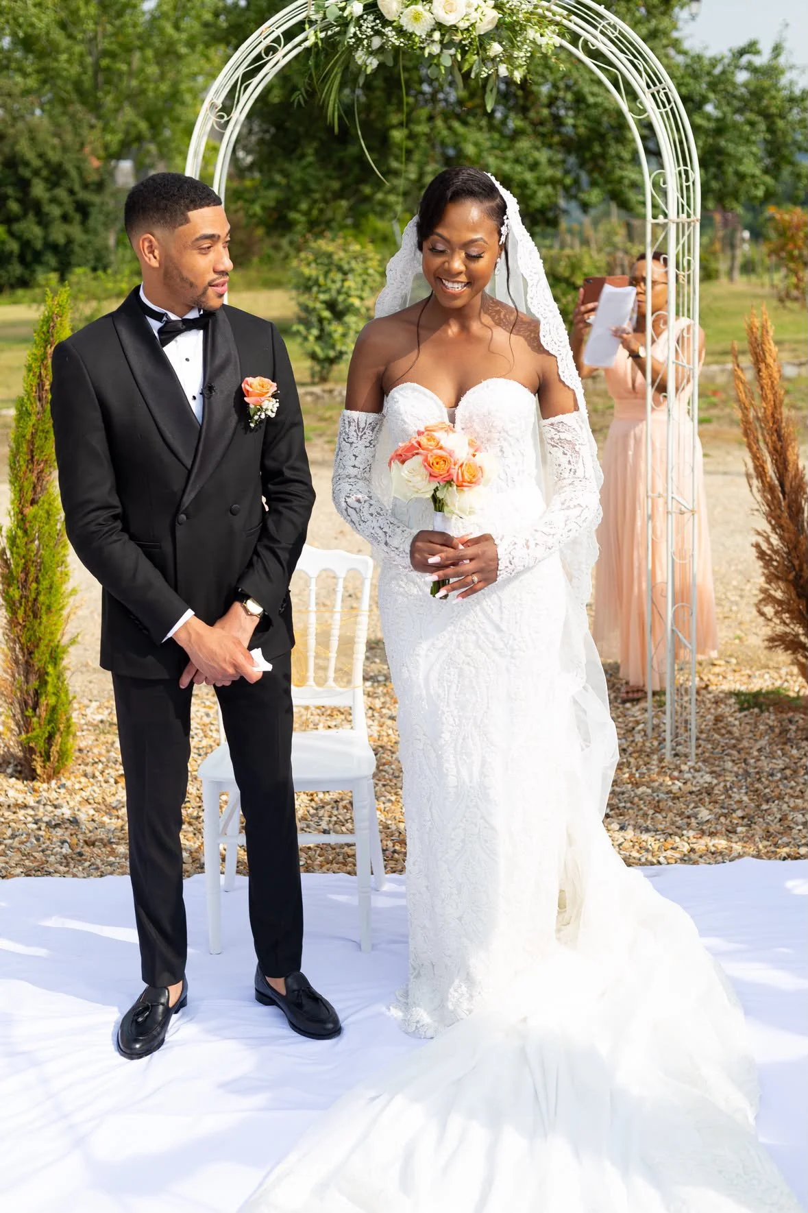 Un couple lors de leur mariage. La femme porte une robe de mariée blanche avec un voile et tient un bouquet de fleurs roses et blanches. L'homme porte un costume noir avec une cravate noire et une fleur de boutonnière. Ils se tiennent devant une arche décorée de fleurs, dans un cadre extérieur avec des arbres en arrière-plan. Une personne en arrière-plan photographie la scène.