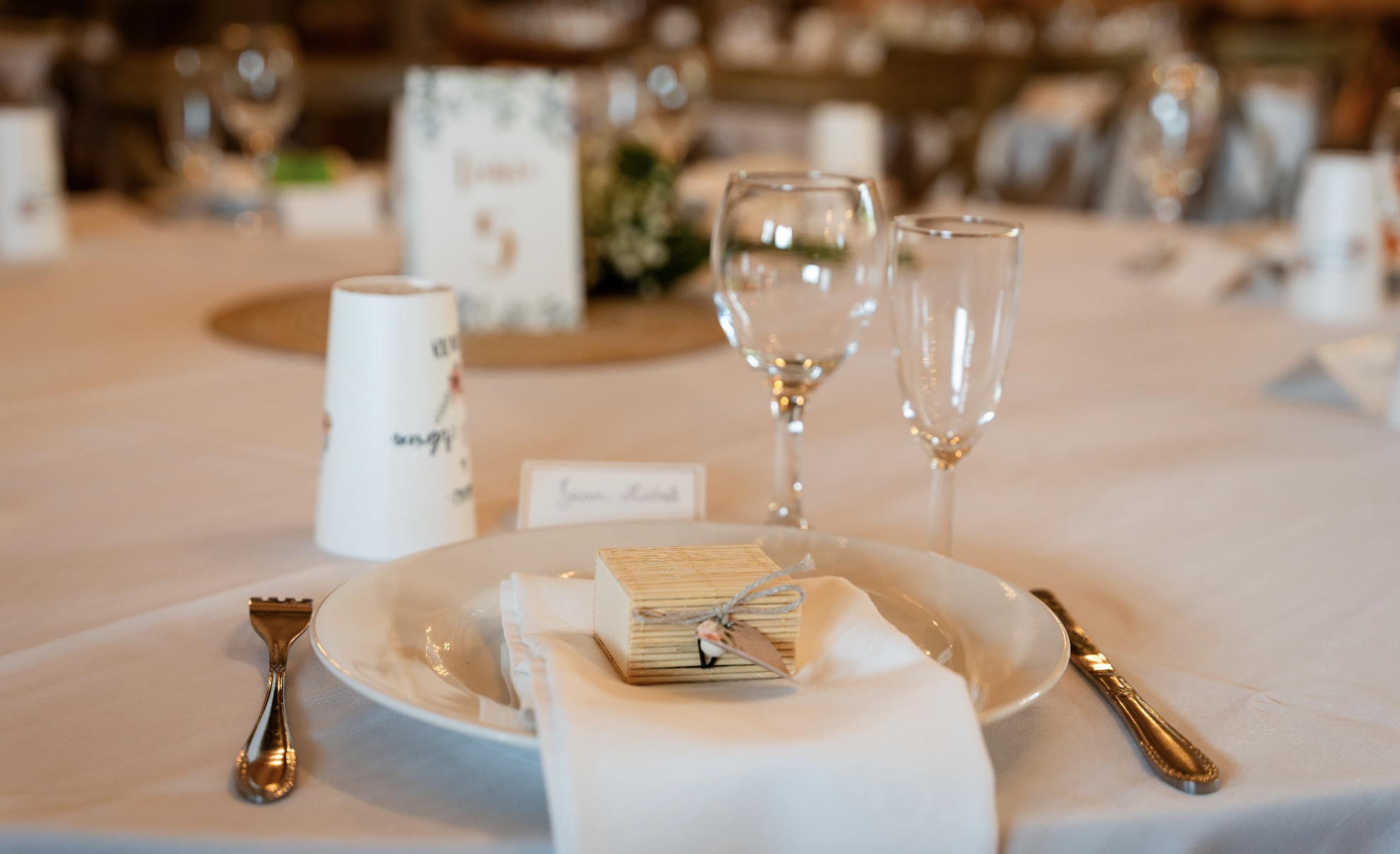 Table de mariage ou de fête avec des verres vides, une assiette avec un petit cadeau emballé et une nappe blanche. En arrière-plan, une décoration florale et une carte de menu ou de place.