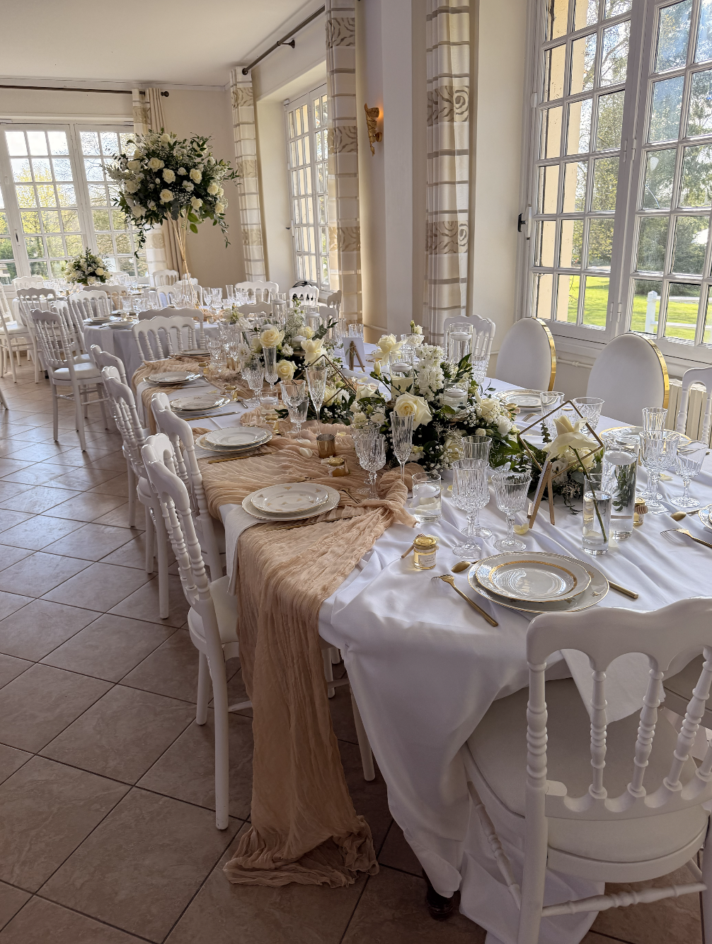 Salle de réception élégante avec une longue table de mariage dressée avec une nappe blanche, des assiettes en porcelaine avec garnitures dorées, des verres en cristal, des bougies et des arrangements floraux blancs, décorée de rideaux beiges avec un 
