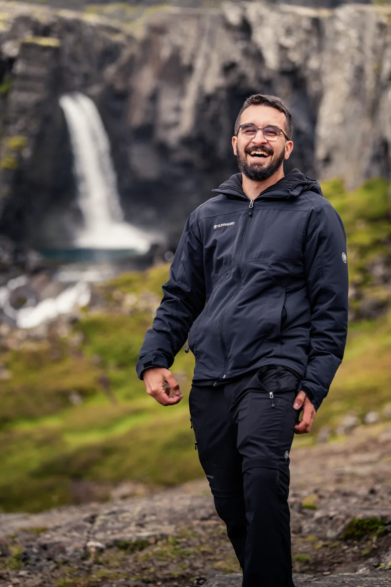 Fotograf Dominik Lüdtke lacht entspannt vor einer Kulisse aus Bergen und einem Wasserfall – euer lockerer Kumpeltyp für Hochzeiten und Familienfotos im Hofer Land.