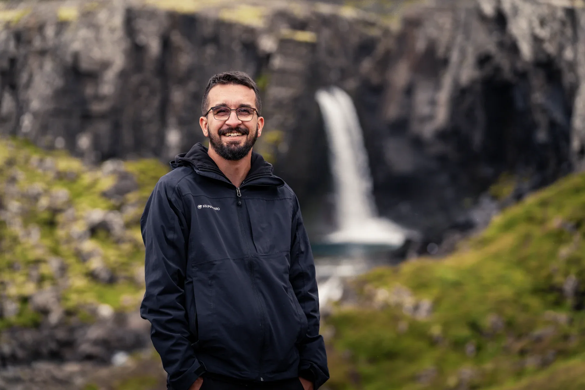 Ein bärtiger Mann mit Brille, Dominik Lüdtke, trägt eine schwarze Funktionsjacke vor einem Wasserfall in einer felsigen, grün bewachsenen isländischen Landschaft.
