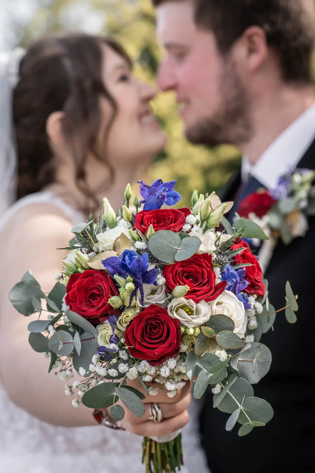 Braut hält einen Blumenstrauß mit roten Rosen, weißen Blumen, blauen Blumen und Eukalyptus, während das Brautpaar sich im Hintergrund liebevoll gegenübersteht. Hochzeit in Oberfranken.