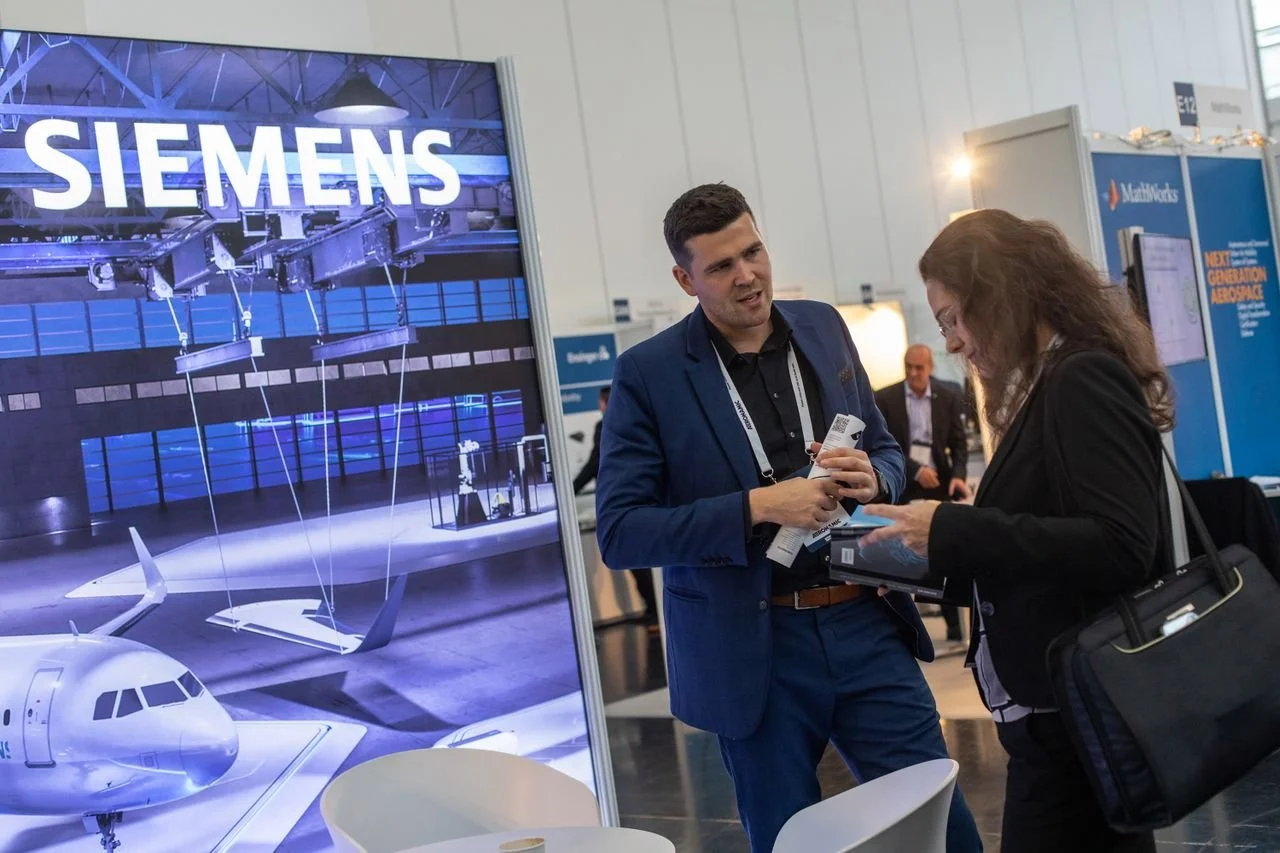 A man in a blue suit and a woman with a black blazer looking at a brochure and talking at an exhibition booth with a Siemens display of aircraft and machinery.