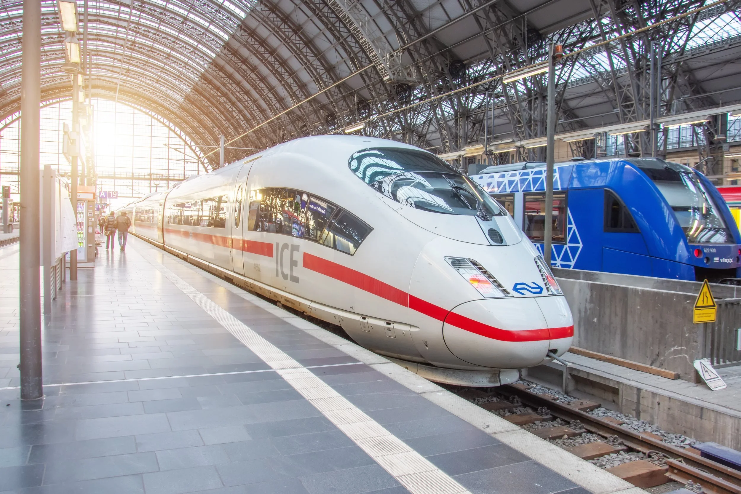 High-speed trains, one white with red stripe and one blue, at a train station with a glass arched roof and some passengers walking nearby.