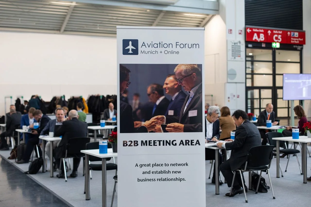 A sign at an aviation forum indicating the B2B meeting area, with people in business attire seated at tables in the background.