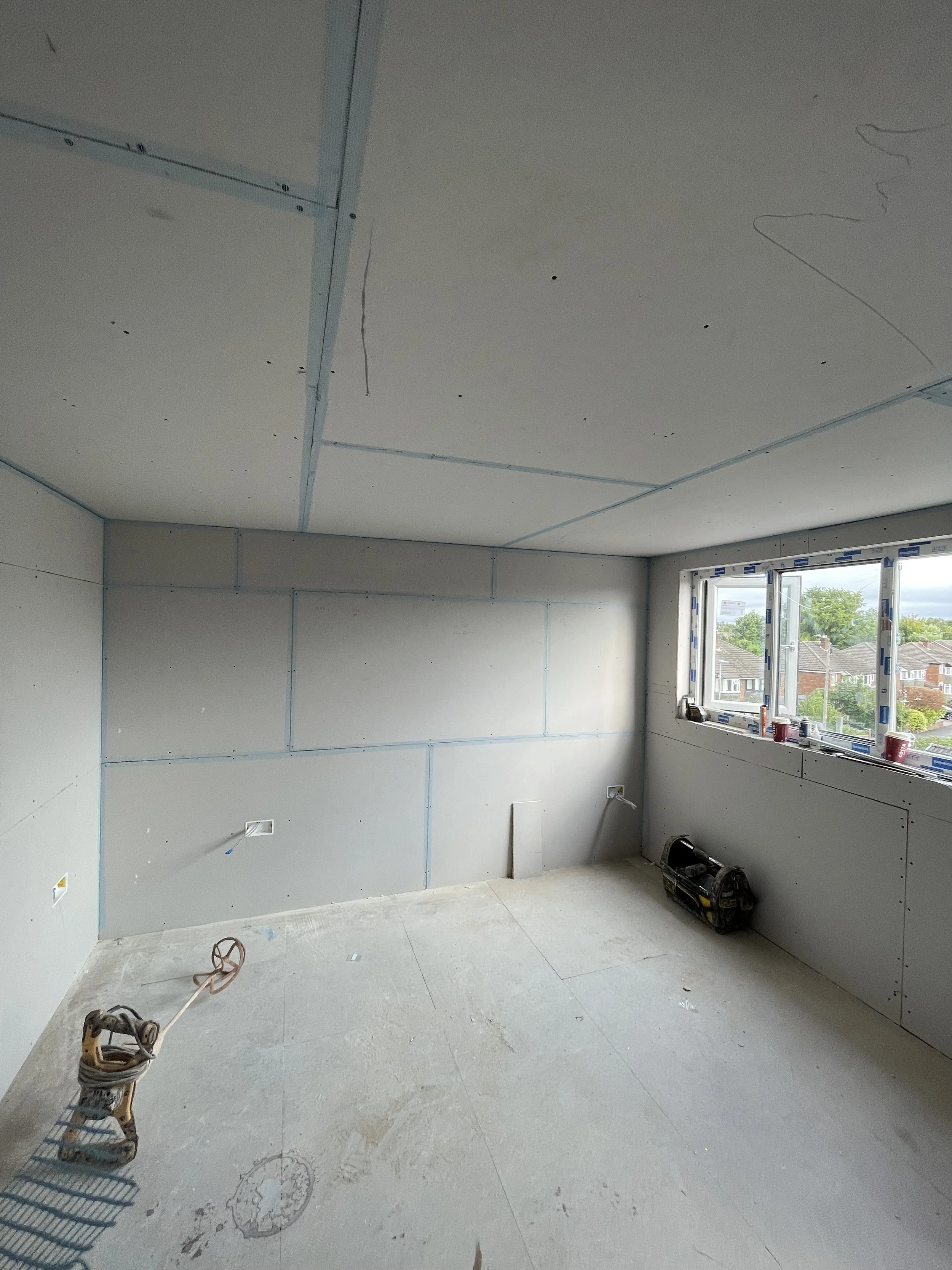 Room under construction with drywall sheets on the walls, a window, construction tools, and wiring on the floor.