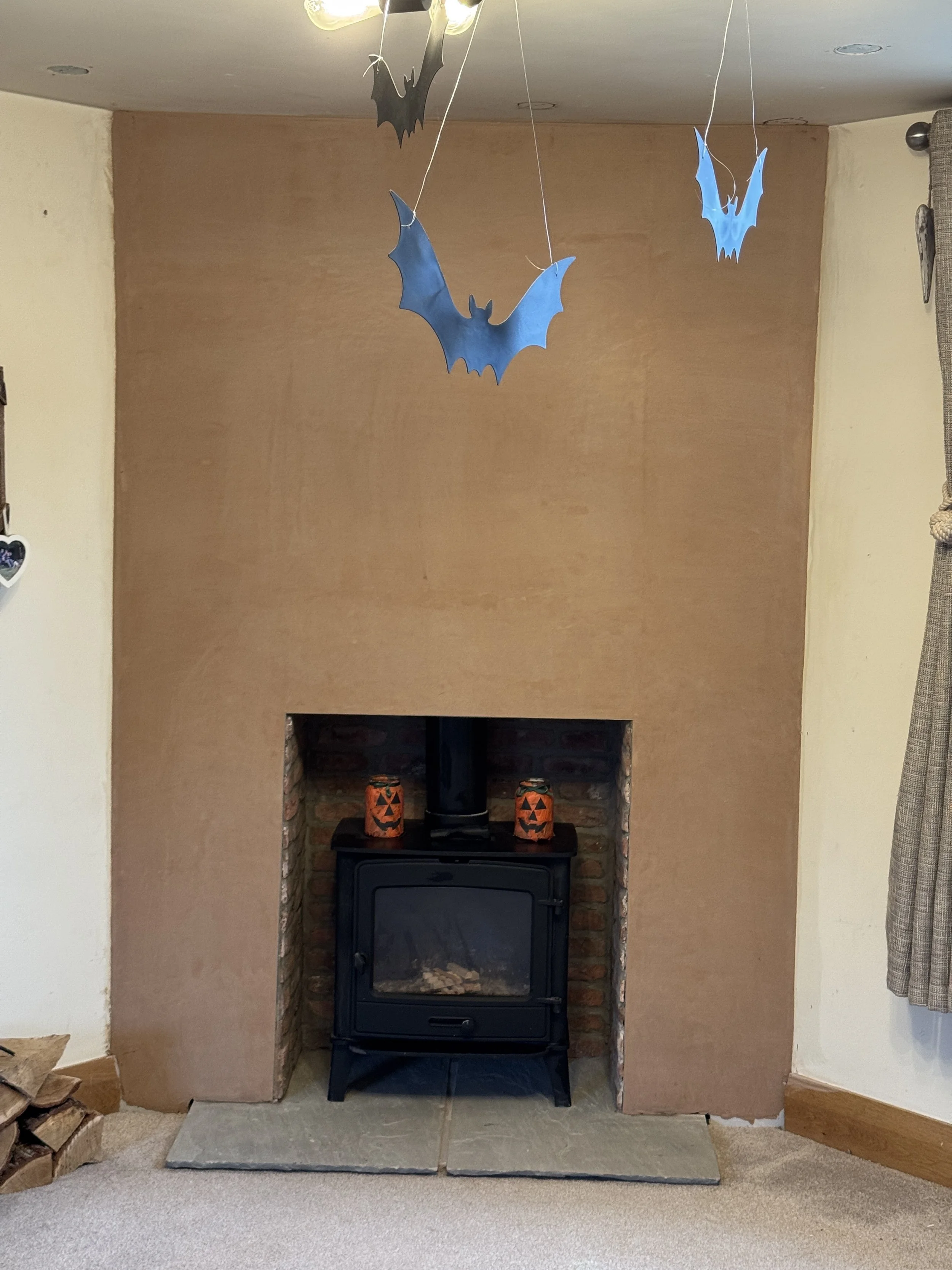 A fireplace with a wood stove inside and a brick surround. Two Halloween-themed lanterns are on top of the stove. The wall around the fireplace is painted a light brown color, and there is a rock hearth beneath it. Cardboard firewood is stacked on th