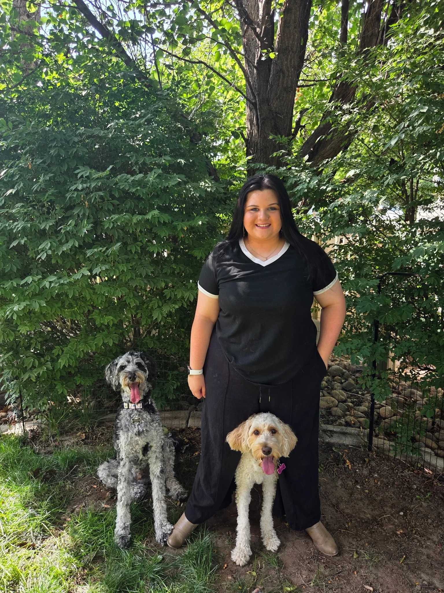 A woman with long black hair smiling standing outdoors in front of green foliage with two dogs, one standing next to her and another sitting in front of her. The woman is wearing a black blouse with white trim and black pants, and tan boots. The dogs are a black and white curly-haired dog and a tan and white dog with a pink tongue hanging out.