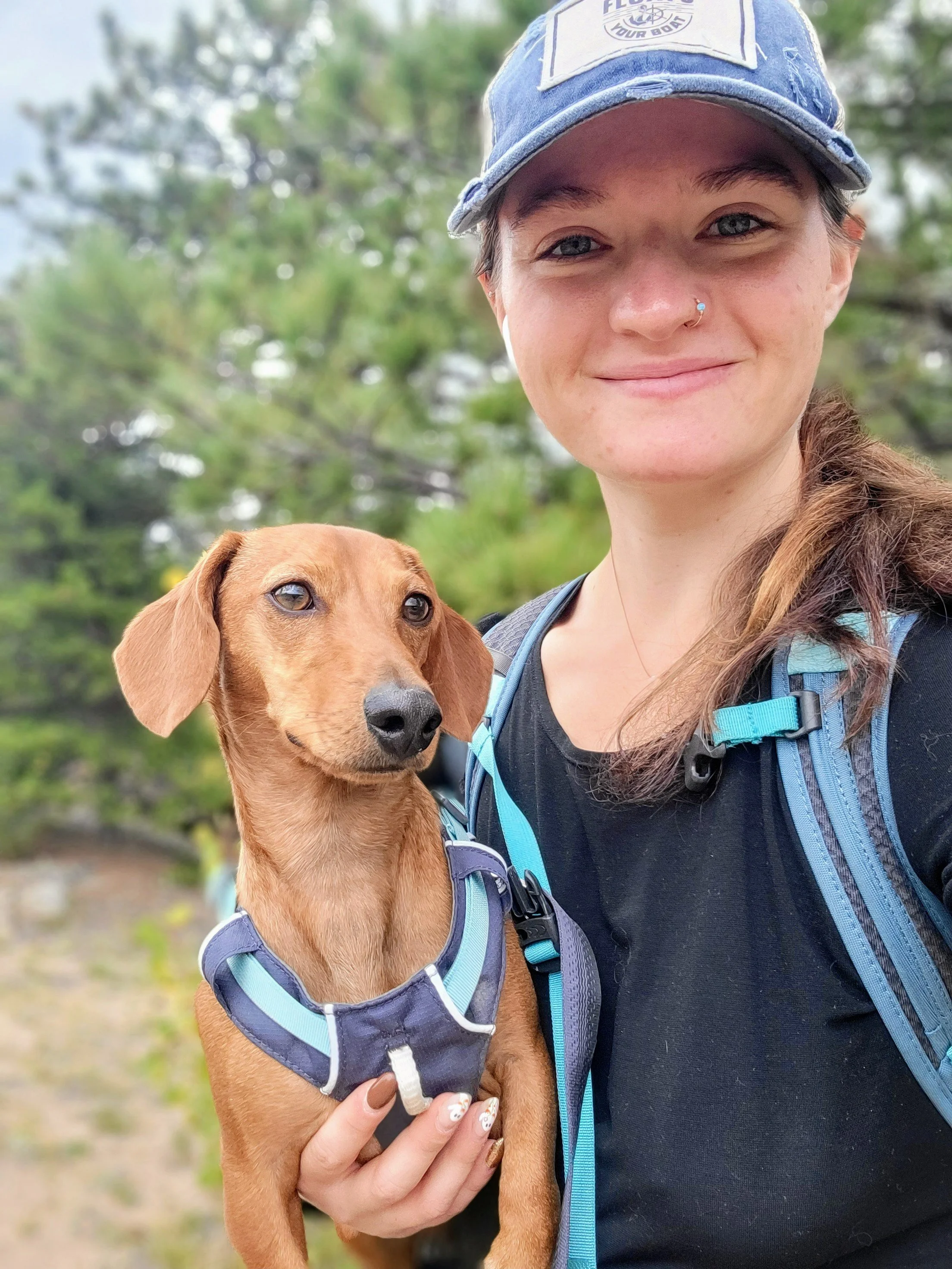 A young woman with a nose piercing, wearing a blue cap, black athletic shirt, and a blue backpack, holding a small brown dog with a blue harness outdoors with green trees in the background.