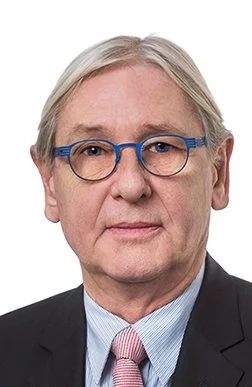 Porträt eines älteren Mannes mit grauen Haaren, Brille mit blauen Rahmen, in formeller Kleidung mit Anzug und Krawatte.