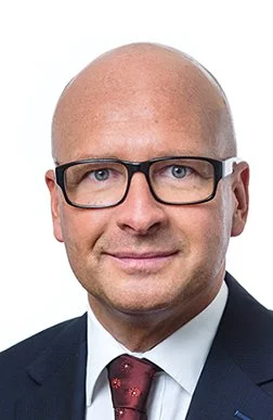 Porträt eines Mannes mit Brille, Anzug und roter Krawatte auf weißem Hintergrund.