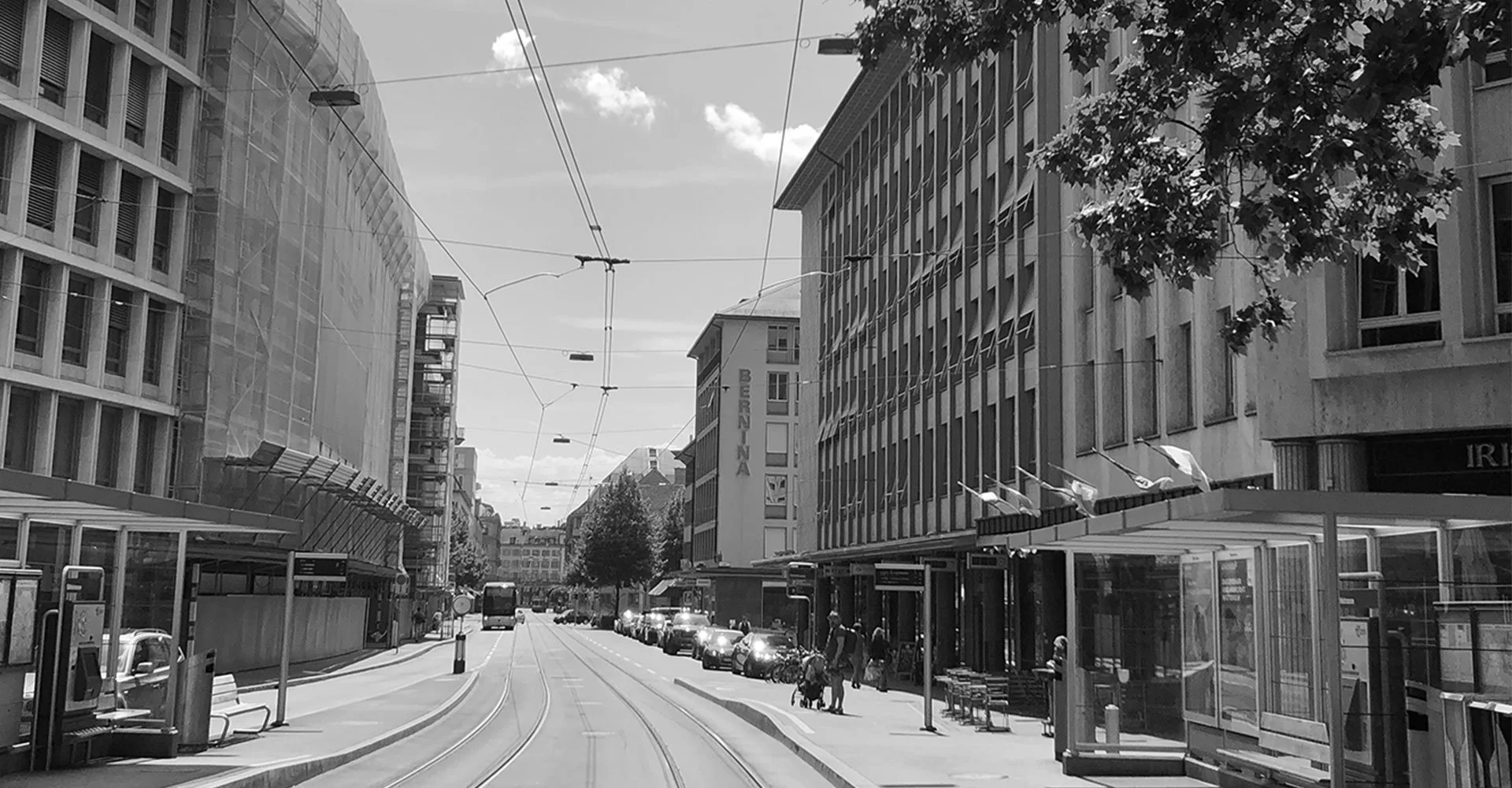 Stadtstraße mit Bahnhof, Tramgleise, modernem Gebäuden und Parkplätzen auf beiden Seiten, einige Menschen und Fahrzeuge, Bäume und eine beschilderte Bushaltestelle.
