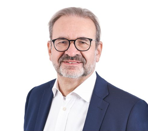 Ein älterer Mann mit Brille, grauem Haar und Bart, trägt einen weißen Hemd und einen dunklen Anzug, lächelt vor weißem Hintergrund.