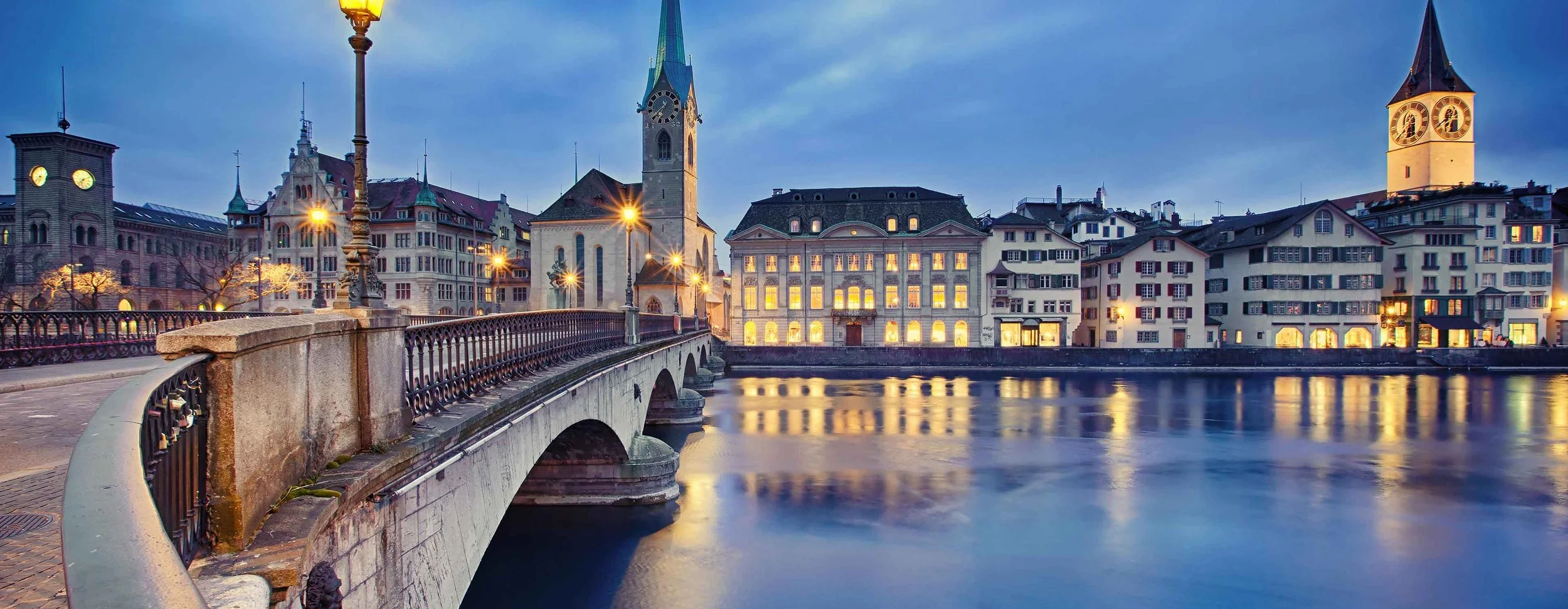 Abendansicht der Stadt Zürich mit alte Brücke über den Fluss, historischen Gebäuden und Kirchtürmen, beleuchtet durch Straßenlaternen bei Dämmerung.