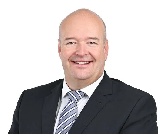 Ein Mann in formeller Kleidung, schwarz Gehrkleid mit Krawatte, lächelnd, vor weißem Hintergrund.