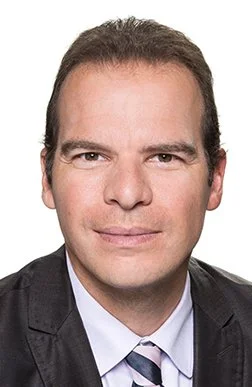 Porträt eines Mannes mit dunklen Haaren in formeller Kleidung, weißes Hemd, dunkler Anzug und Krawatte, vor weißem Hintergrund.