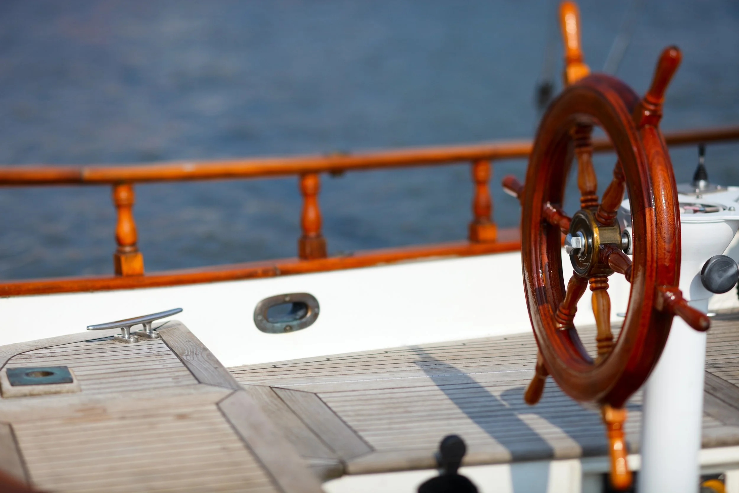Das Bild zeigt das Steuer eines Segelboots, bestehend aus einem großen Holzrudder und einer Steuerrad. Im Hintergrund ist Wasser sichtbar.