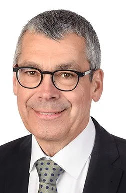 Porträt eines smiling Mannes mit Brille, grau Haare, in einem Anzug mit Krawatte, vor weißem Hintergrund.